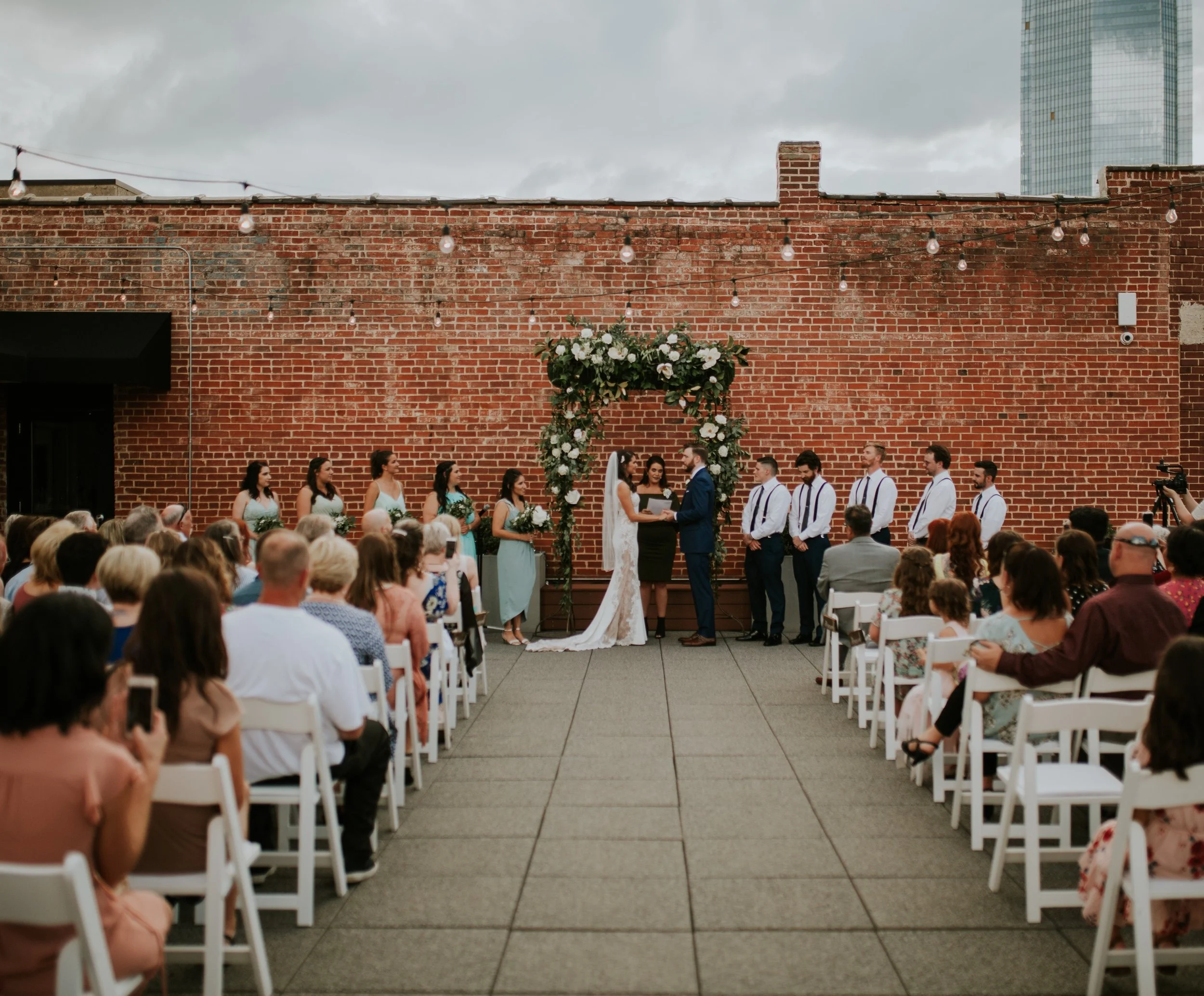 A wedding ceremony taking place outdoors on a rooftop with a brick wall background and string lights overhead. The bride and groom are at the altar with the officiant, surrounded by guests seated in white chairs. Bridesmaids and groomsmen stand on ei