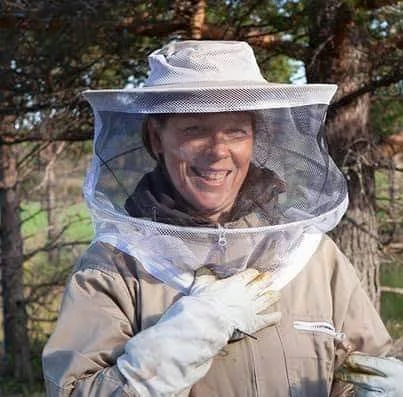 HonnIngunn iført birøkterdrakt, smilende, ute i naturen ved bigården med trær i bakgrunnen.