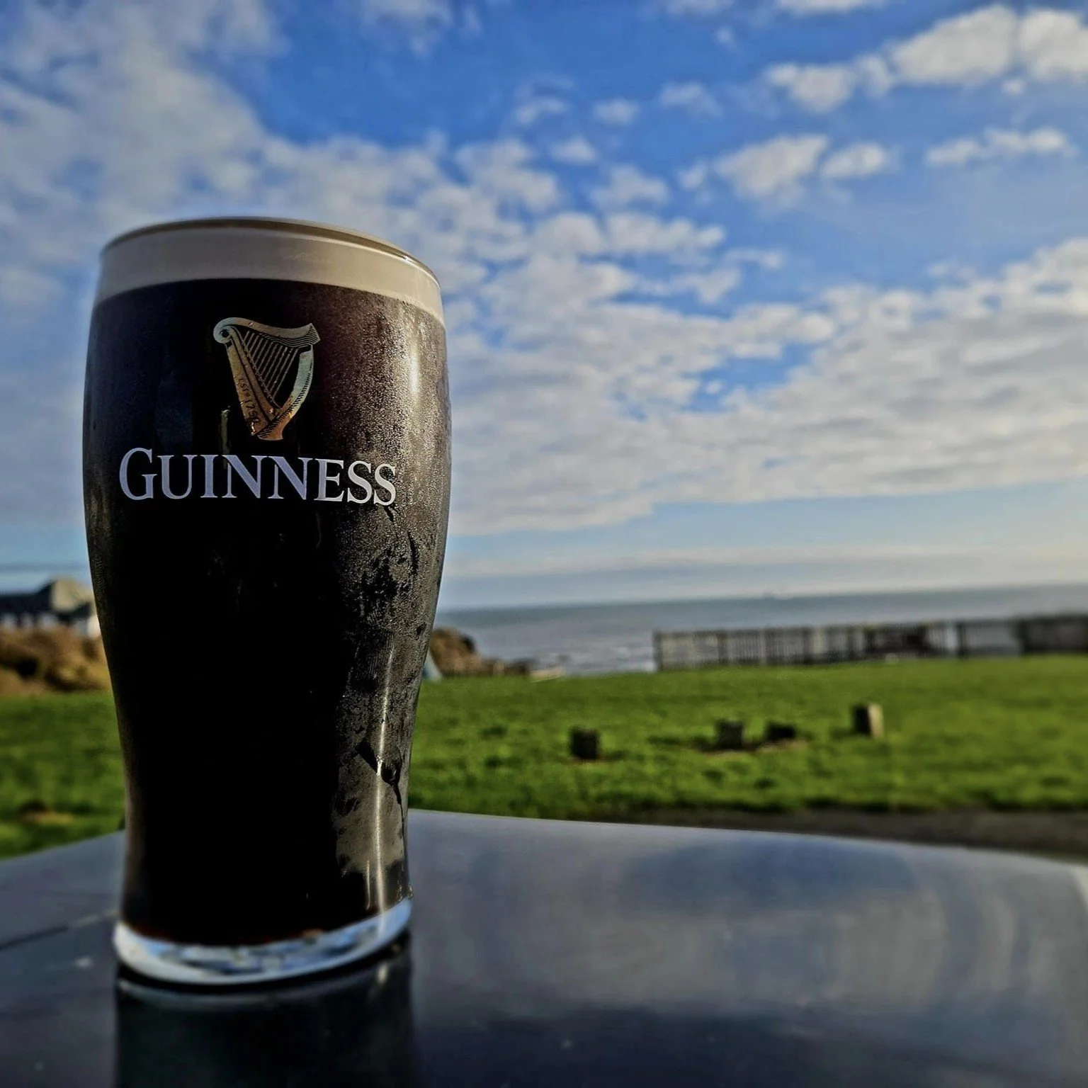 A glass of Guinness beer with a view of an ocean, sky with scattered clouds, a grassy area, and a pier in the background.