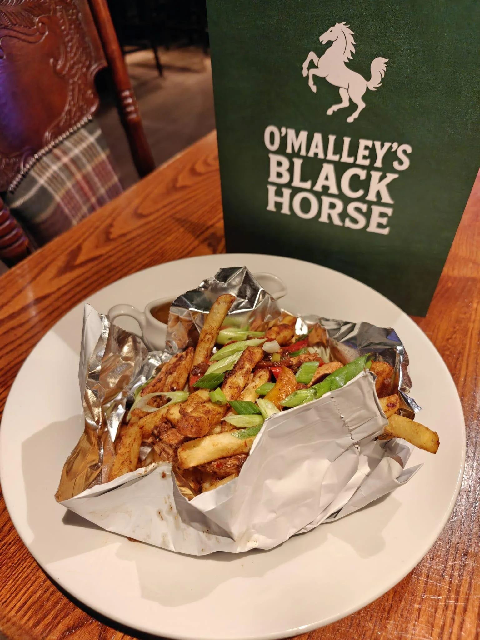 A plate of French fries with fried chicken pieces and chopped green onions, served with a dipping sauce, on a wooden table with a green O'Malley's Black Horse menu beside it.