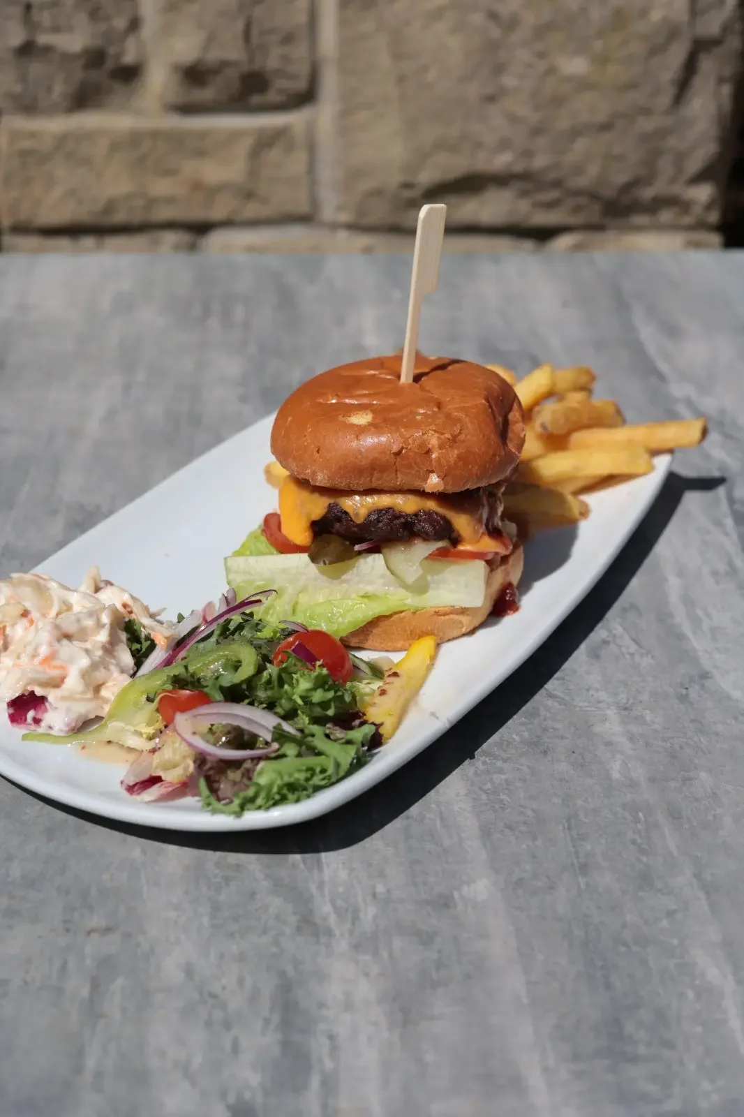 Cheeseburger with lettuce, tomato, pickles, and cheese, served with French fries and a side salad on a white platter.