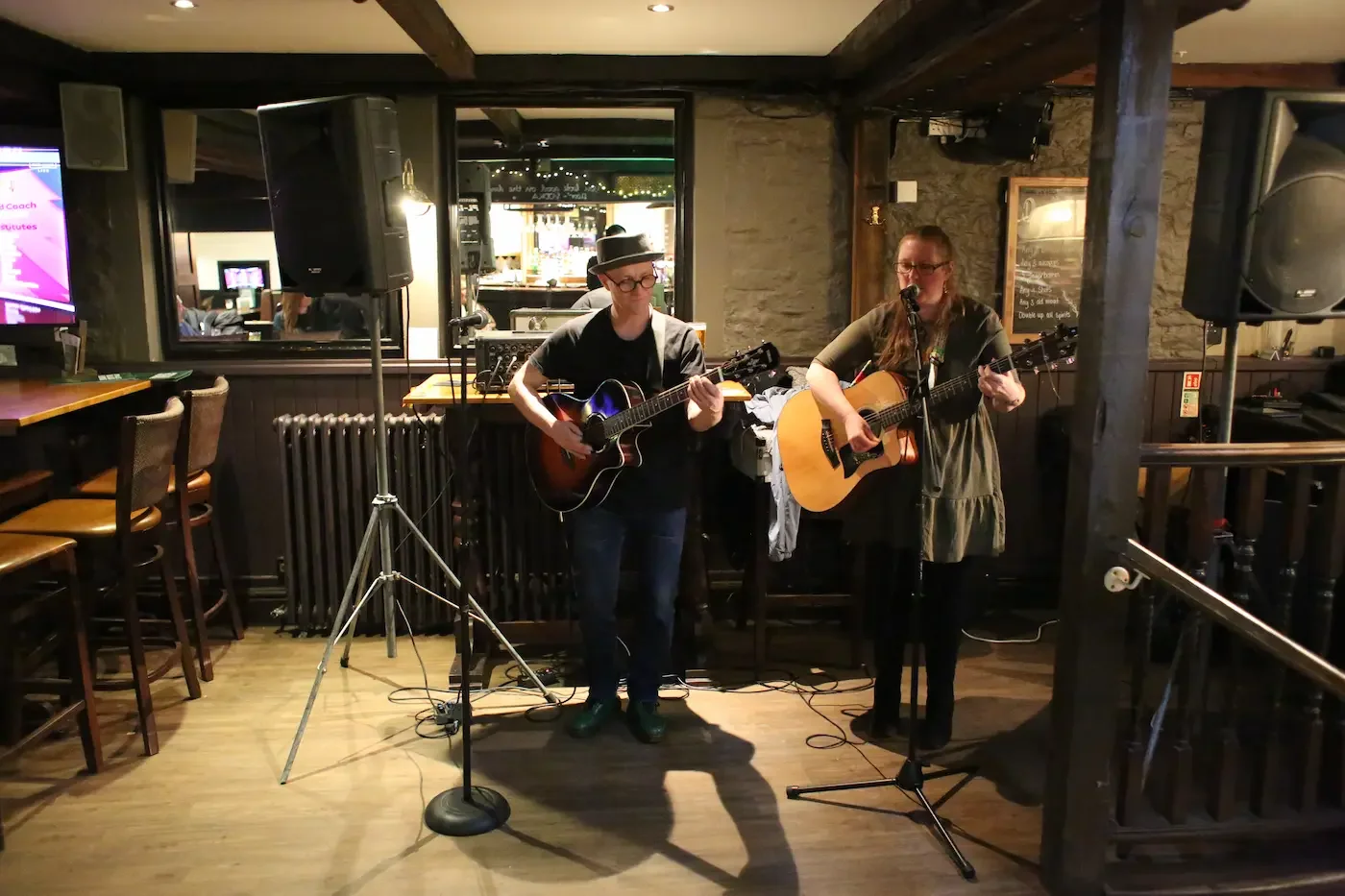 Two musicians playing guitars and singing into microphones in a cozy indoor setting, with speakers and sound equipment, wooden beams, and bar seating in the background.