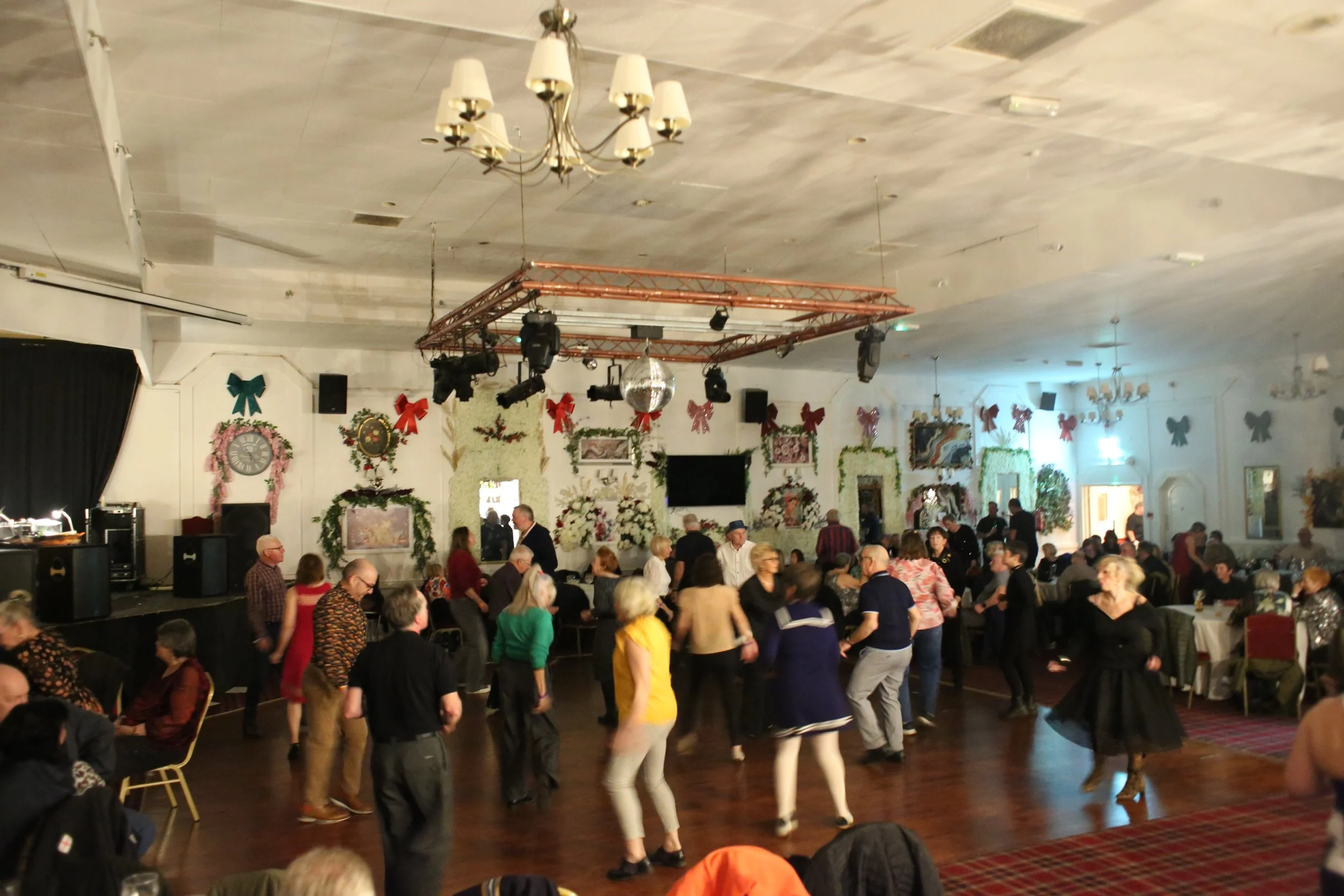People dancing on a wooden floor in a decorated banquet hall with Christmas decorations, wreaths, bows, and flowers, while others are seated at tables. A stage with speakers and lights is visible.