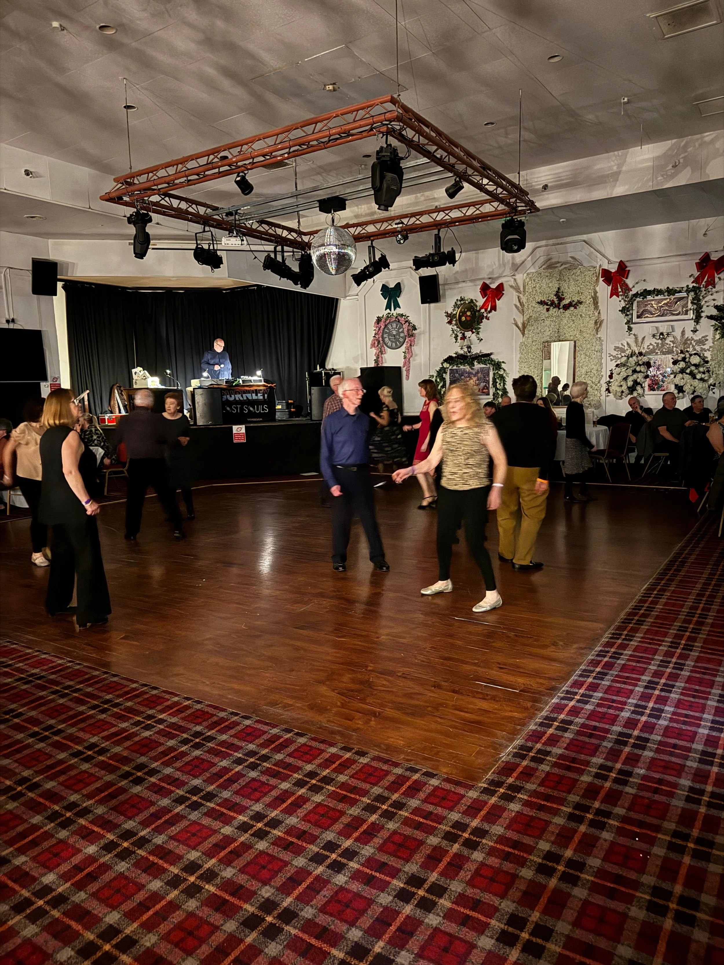 People dancing on a wooden dance floor at a holiday event with Christmas decorations and a DJ on stage.