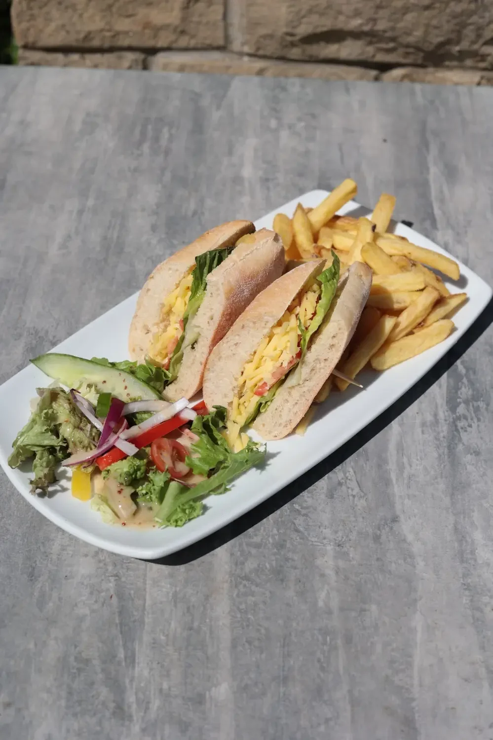 A white rectangular plate with a sandwich containing lettuce, tomato, cheese, and deli meat, served with a side of French fries and a small side salad with mixed greens, cucumber, tomato, and onion.