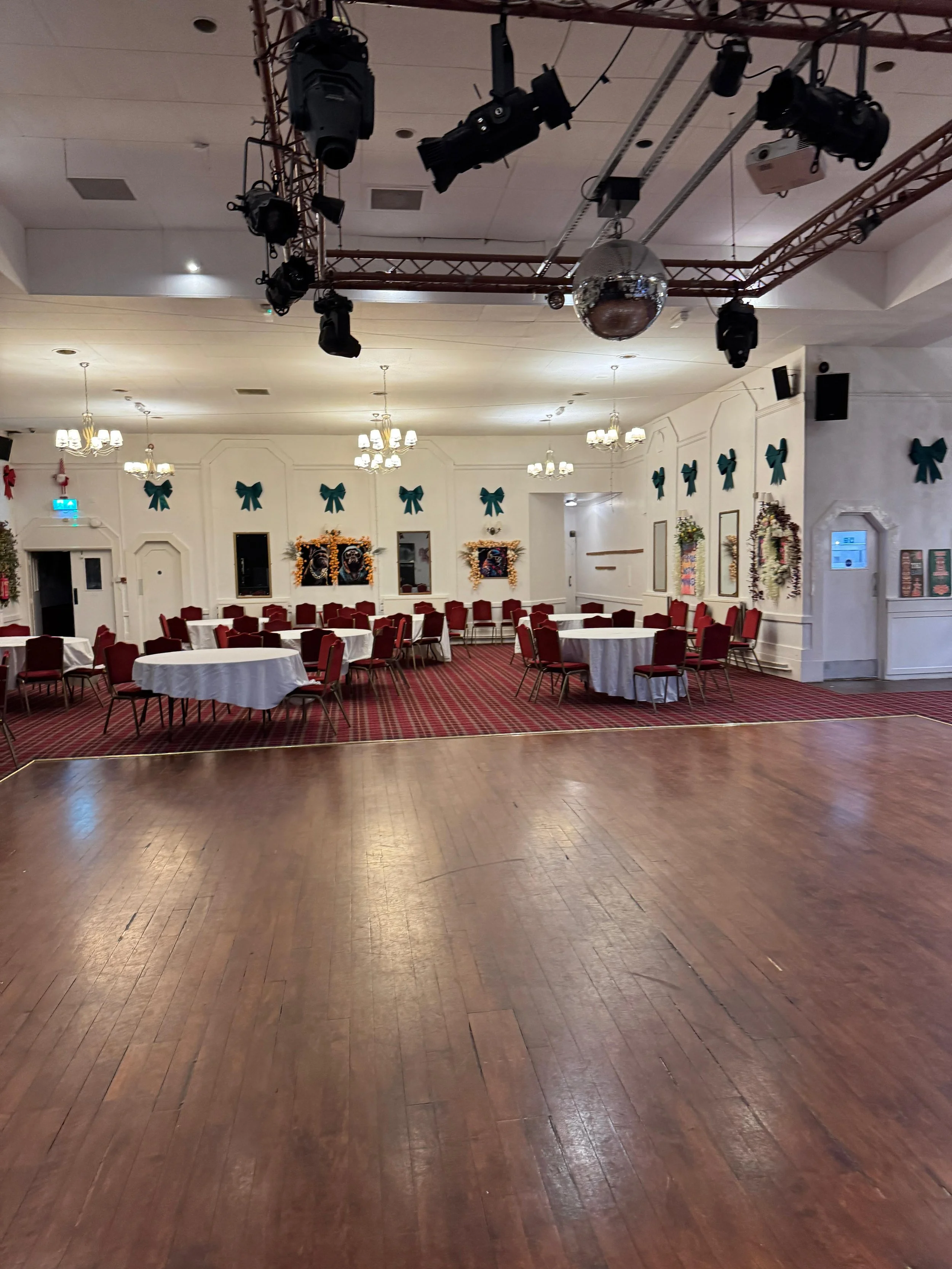 Event hall decorated with green bows, wreaths, and framed pictures, set up with round tables and red chairs, with stage lighting and a disco ball overhead.