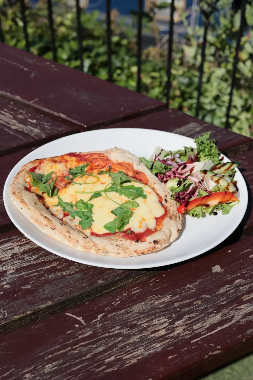 A plate with a personal-sized cheese pizza with fresh basil on top and a side salad with mixed greens, cucumbers, and tomatoes. The plate is on a weathered wooden outdoor table.