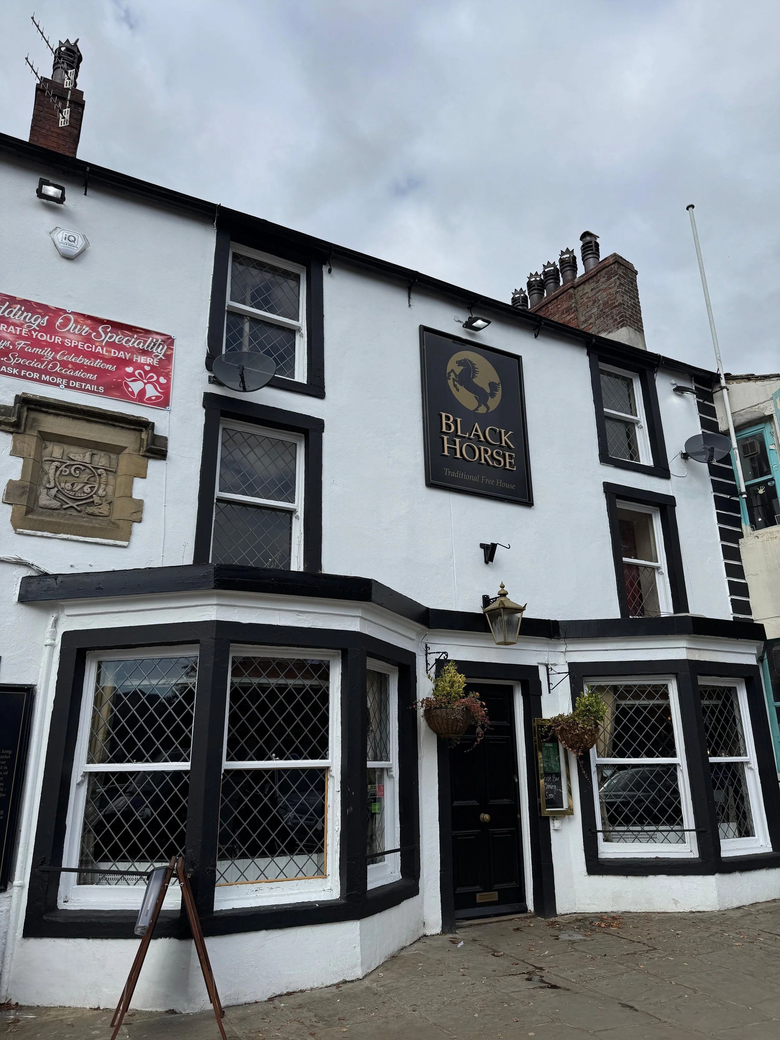 Exterior of a white building with black trim, featuring large bay windows with decorative security grilles, a black door with hanging flower baskets, and a sign that reads 'Black Horse Traditional Free House', with a logo of a black horse.