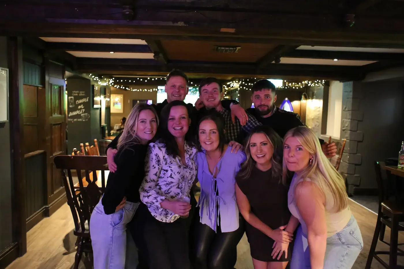 A group of eight friends smiling and posing together in a cozy pub or restaurant, with string lights and a TV in the background.