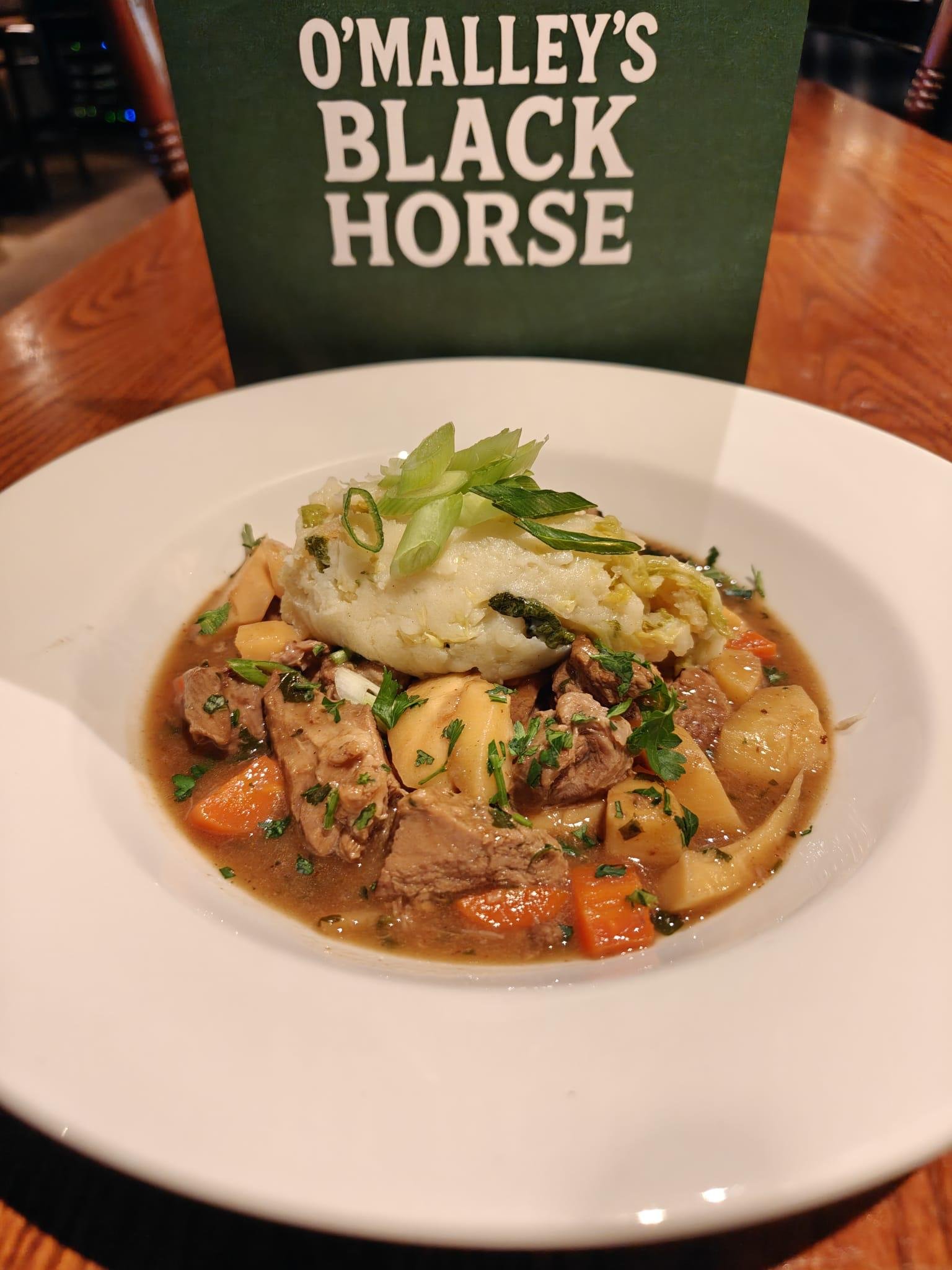 A bowl of stew with chunks of beef, carrots, potatoes, and herbs, topped with mashed potatoes and garnished with green onions on a wooden table, with a sign behind it reading "O'Malley's Black Horse."