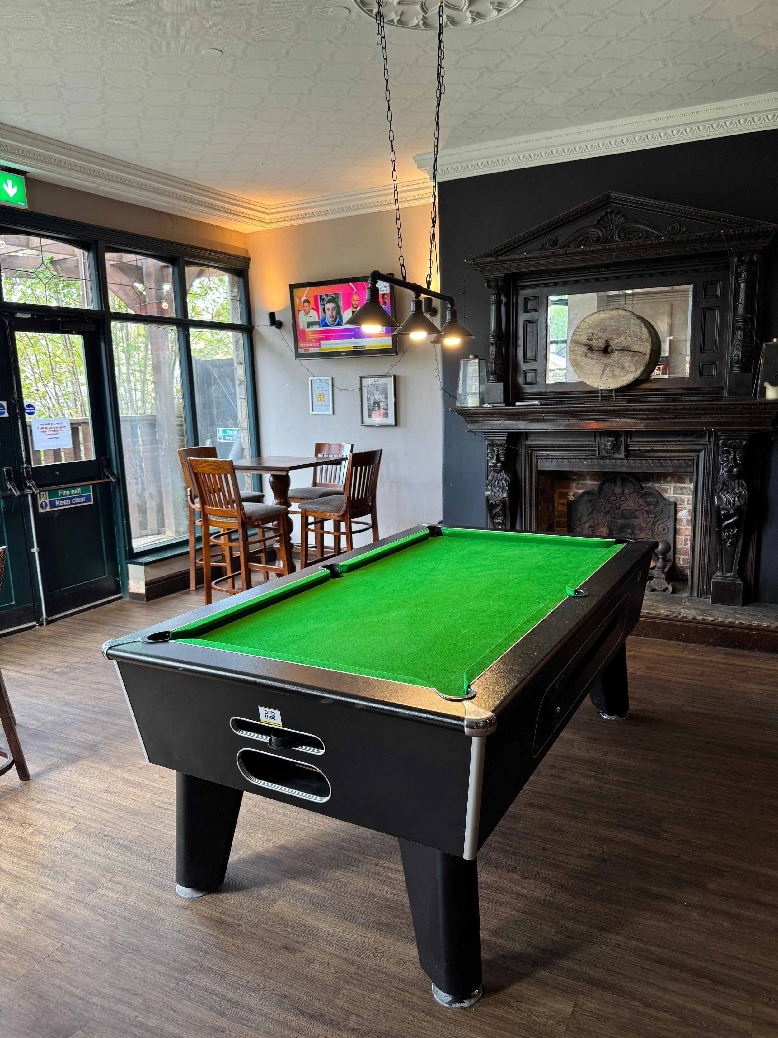 A black pool table with green felt in a cozy room with wooden flooring, a black ornate fireplace, wall-mounted TV, and large windows with outdoor greenery.