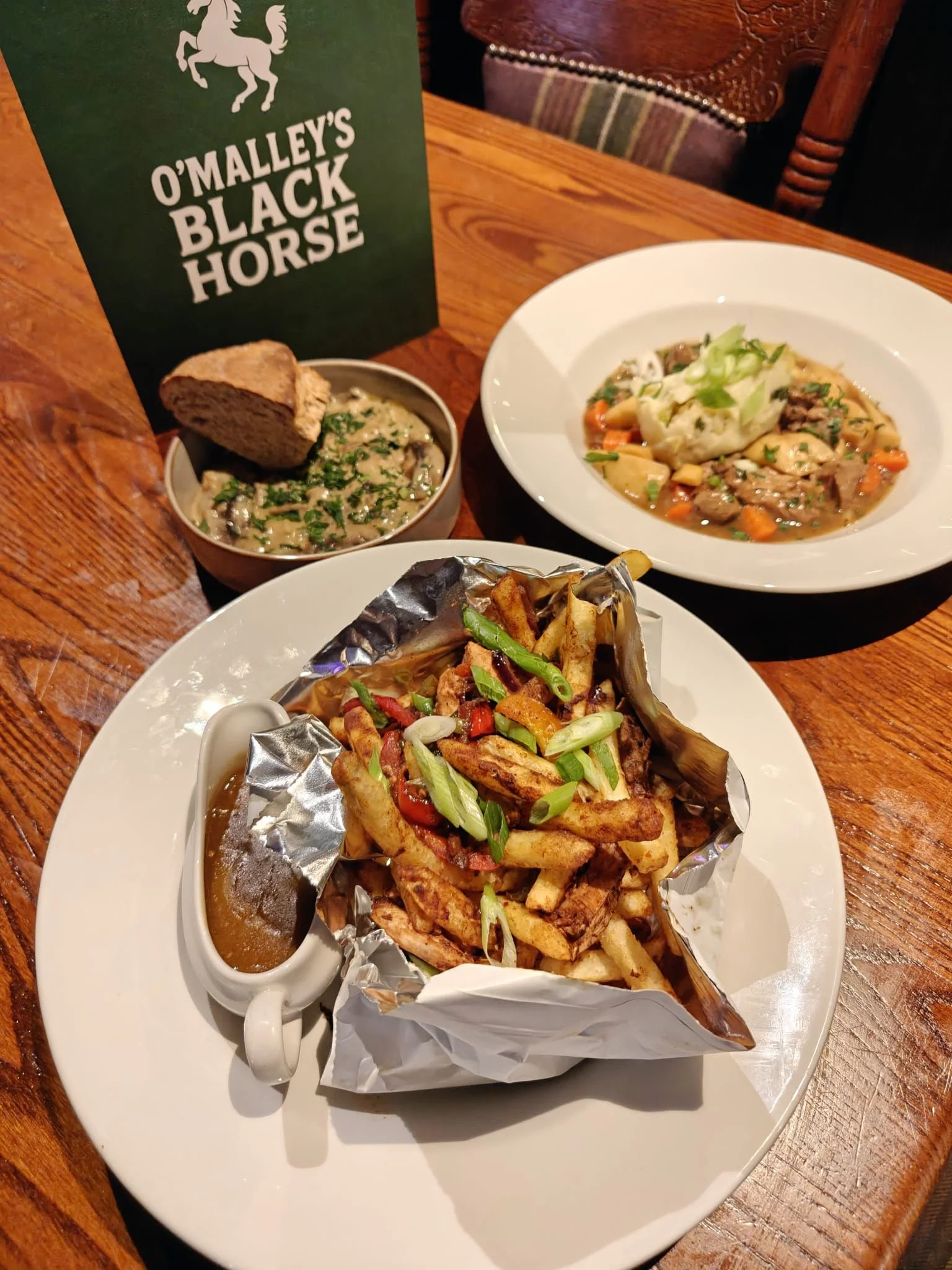 Plate of French fries topped with green onions, served with a side of gravy. In the background, there are two bowls of stew or gravy, one with a piece of bread, on a wooden table with a green O'Malley's Black Horse sign.