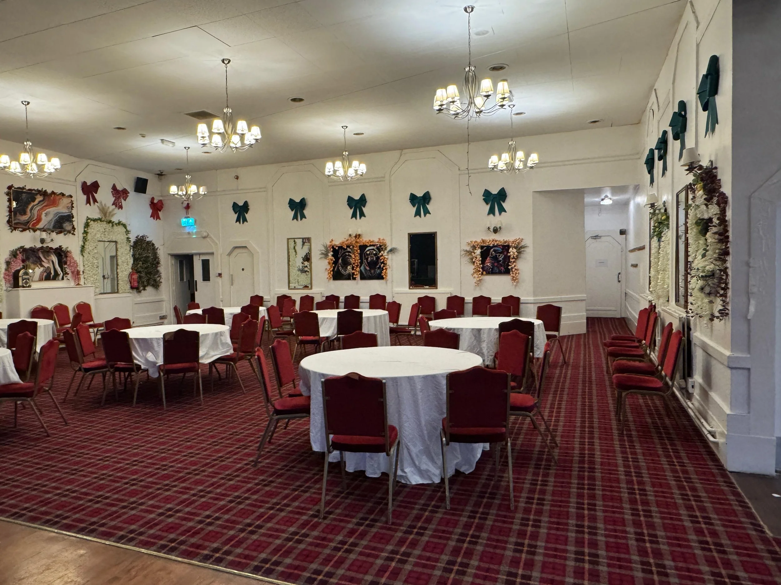 A banquet hall decorated for a celebration with red bows on the wall, floral arrangements, and chandeliers. Several round tables with white tablecloths and red chairs are arranged in the room.