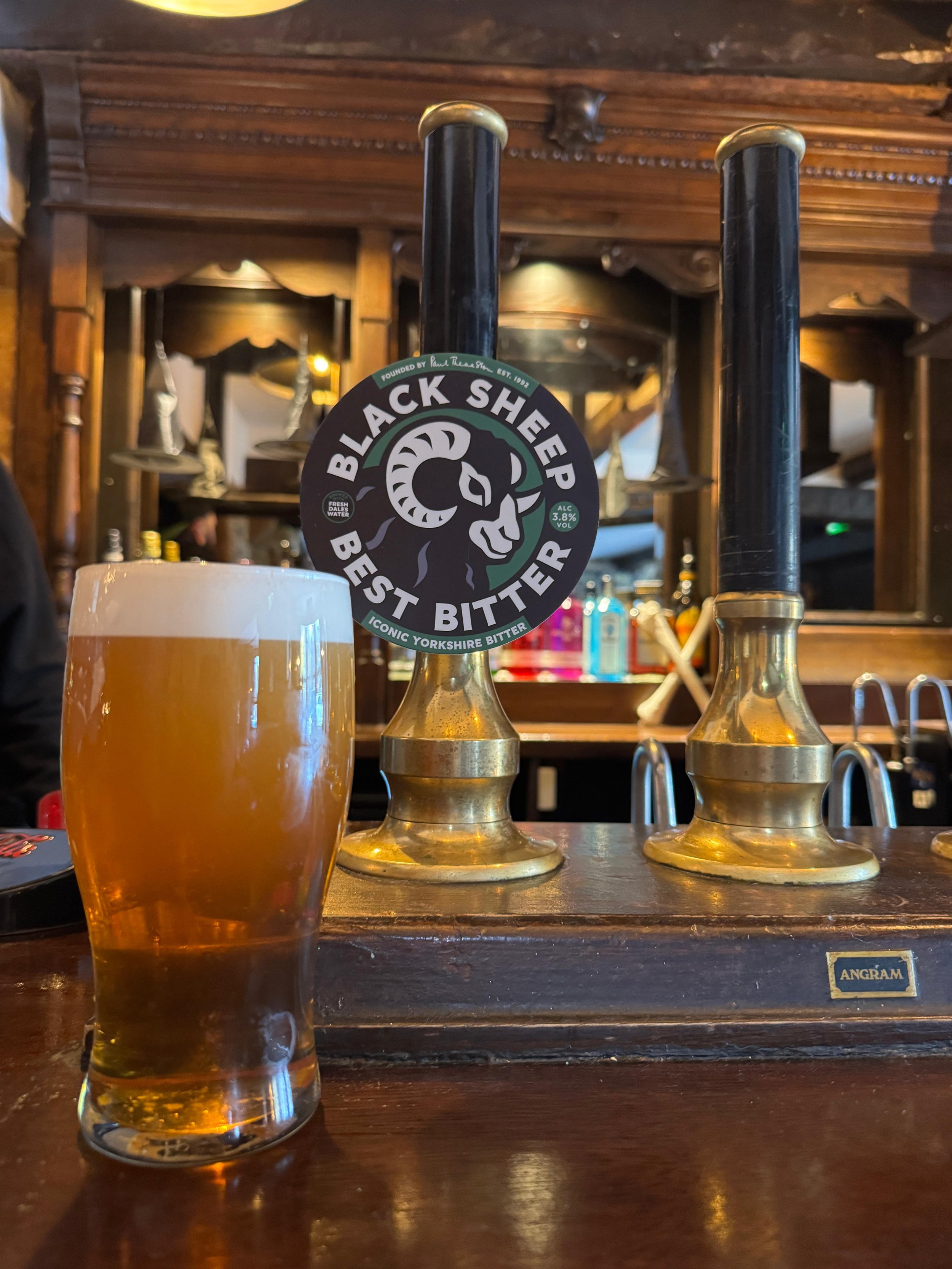 A glass of light amber beer with a foamy head on a wooden bar counter, with two beer taps labeled 'Black Sheep Best Bitter' behind it in a pub setting.