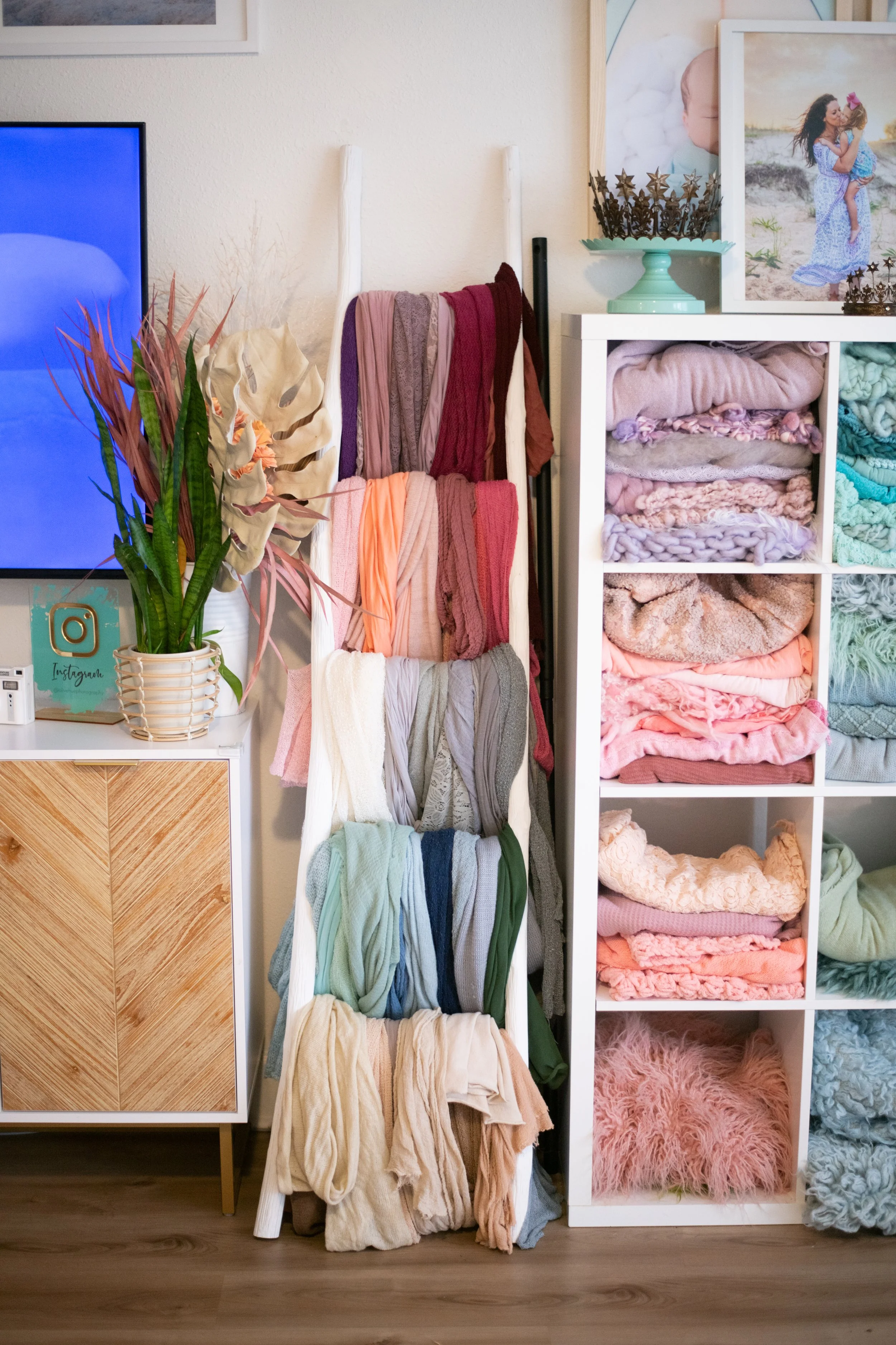 A room corner with a white shelf filled with folded and draped blankets, a small wooden cabinet, and a display of colorful scarves hanging on a white stand. There is a potted plant with long green and reddish leaves on top of the cabinet, and framed