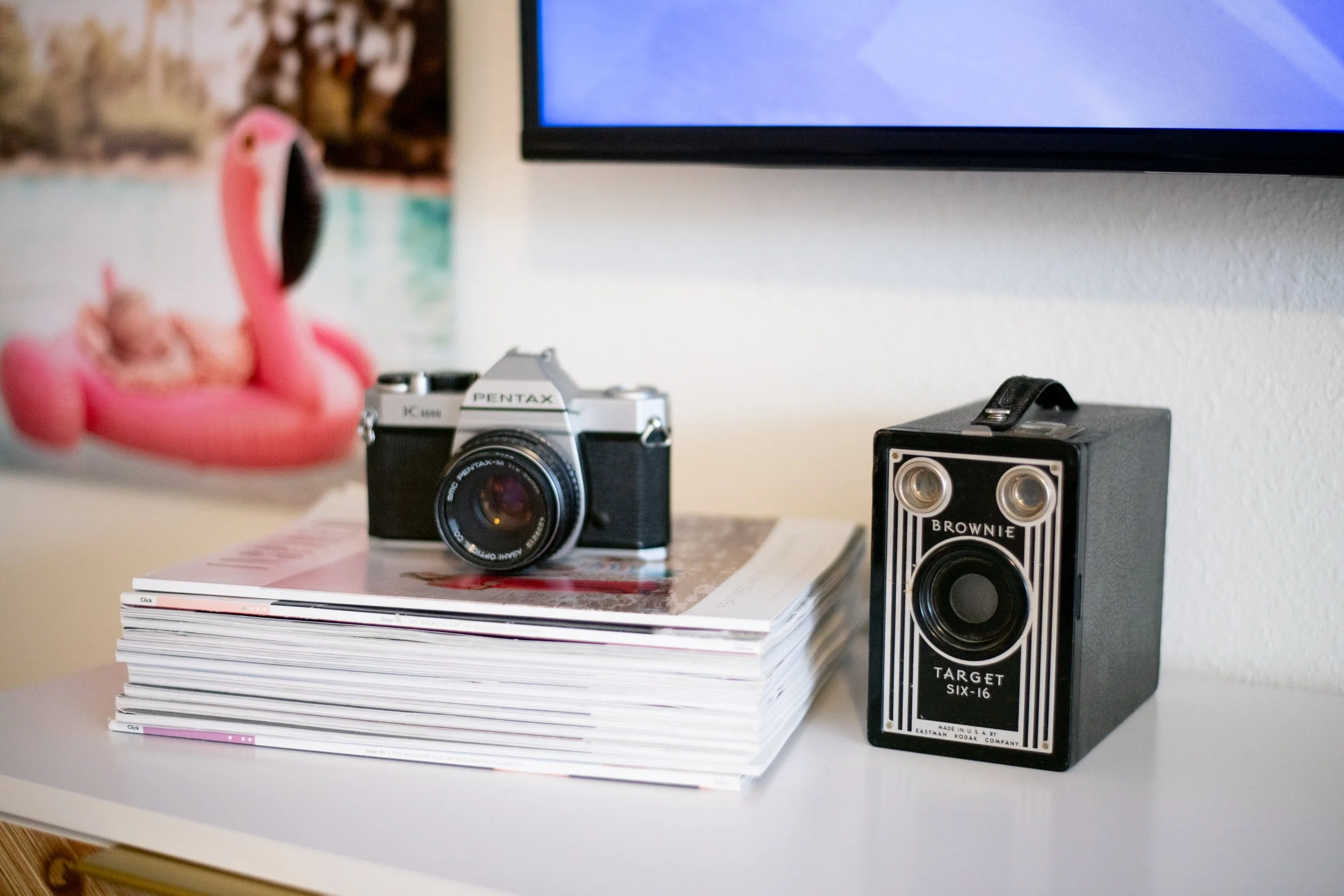 A vintage Pentax camera placed on a stack of magazines, with a vintage Brownie Target Six-16 camera to the right, in front of a white wall and a partial view of a television screen. A pink flamingo-shaped decoration is partially visible on the left s