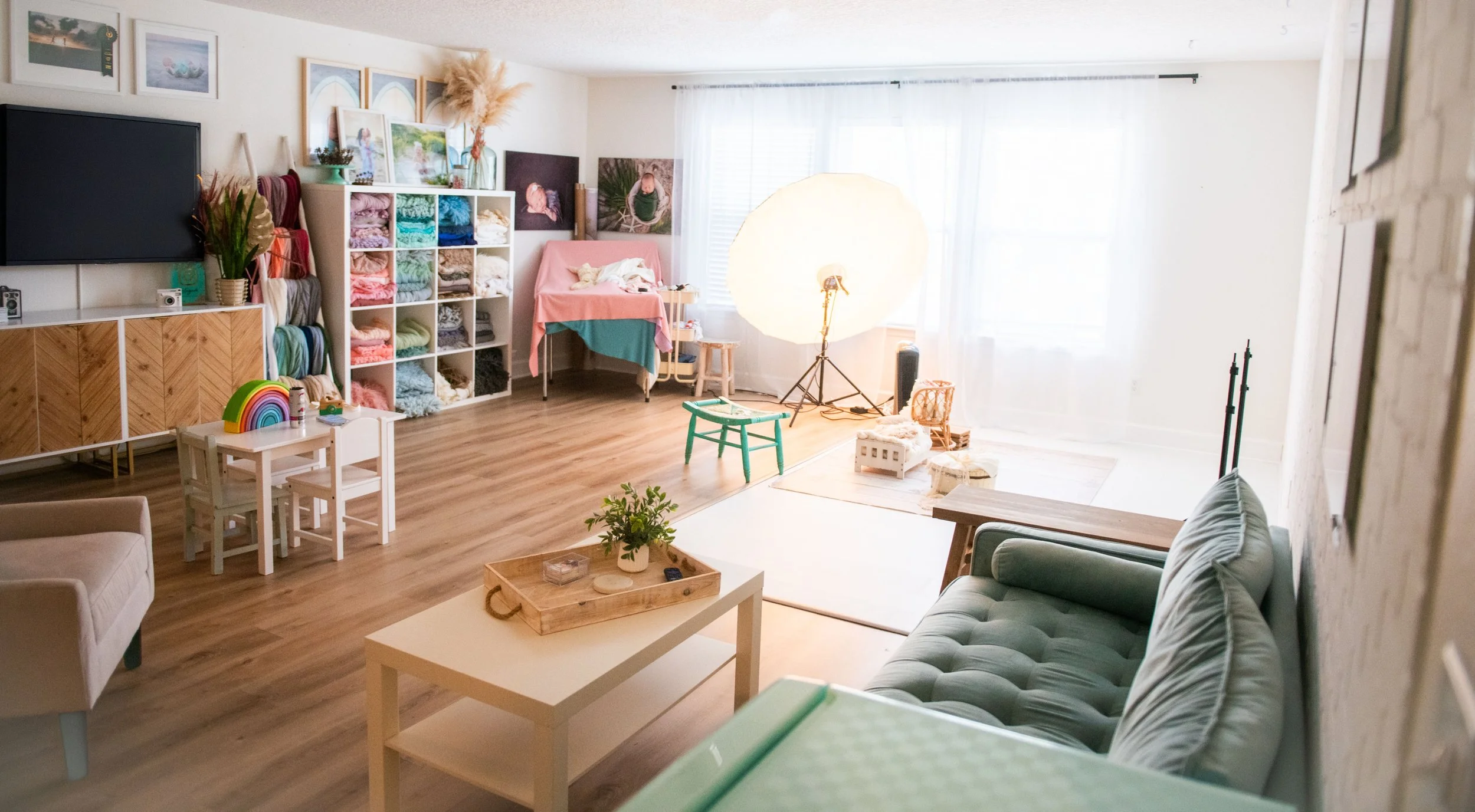 Bright living room with a green sofa, wooden flooring, a white coffee table with a plant, a small children's table with three chairs, a wall-mounted TV, colorful yarn and fabric storage, framed artwork, and a photography studio light with background,
