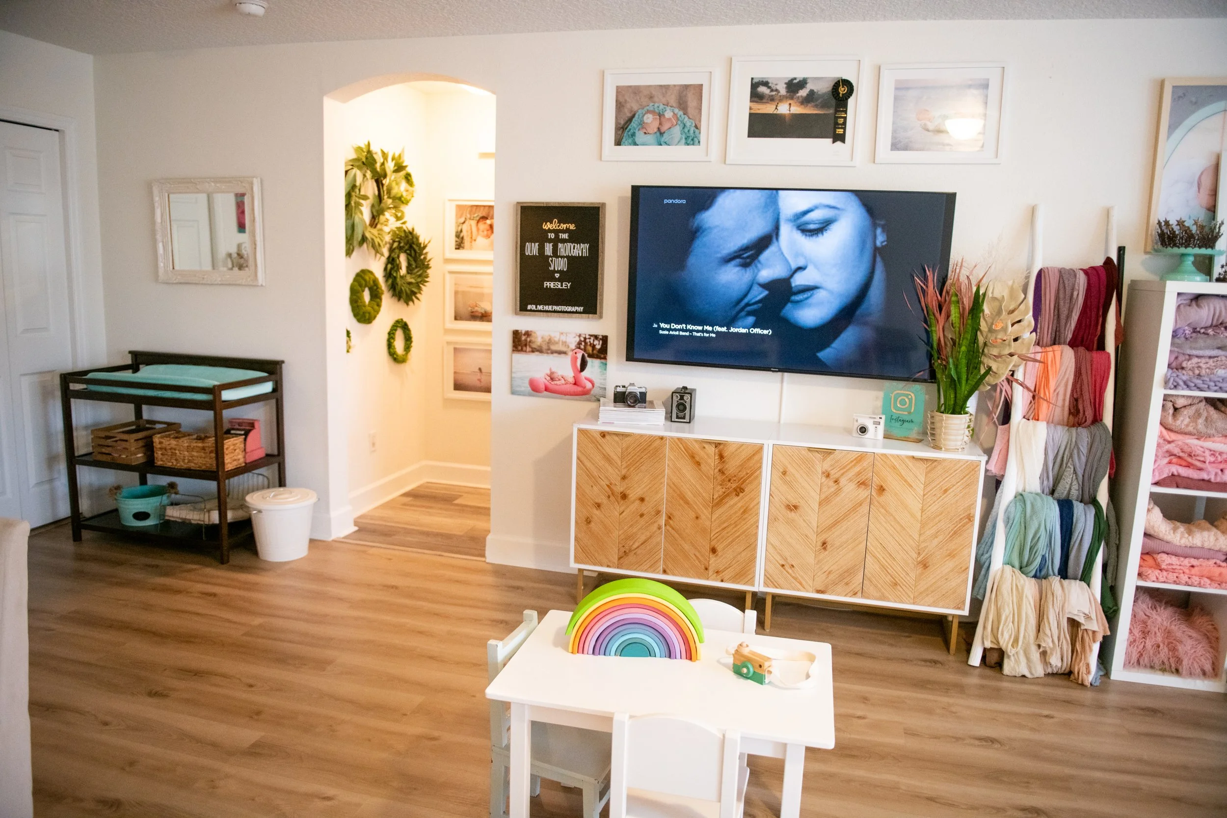 Living room with a television on the wall showing a black and white scene from a movie, a photo display above, a small table with a rainbow toy, and a storage cart with baskets. There are shelves with folded blankets and a plant with colorful leaves.