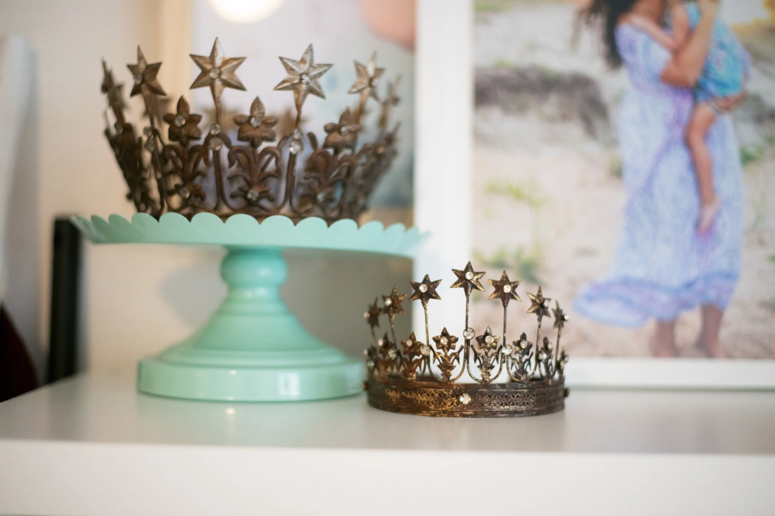 Two decorative crowns with star and flower designs, one on a mint green stand and one on a table, with a blurred beach photo of a girl in a light blue dress in the background.