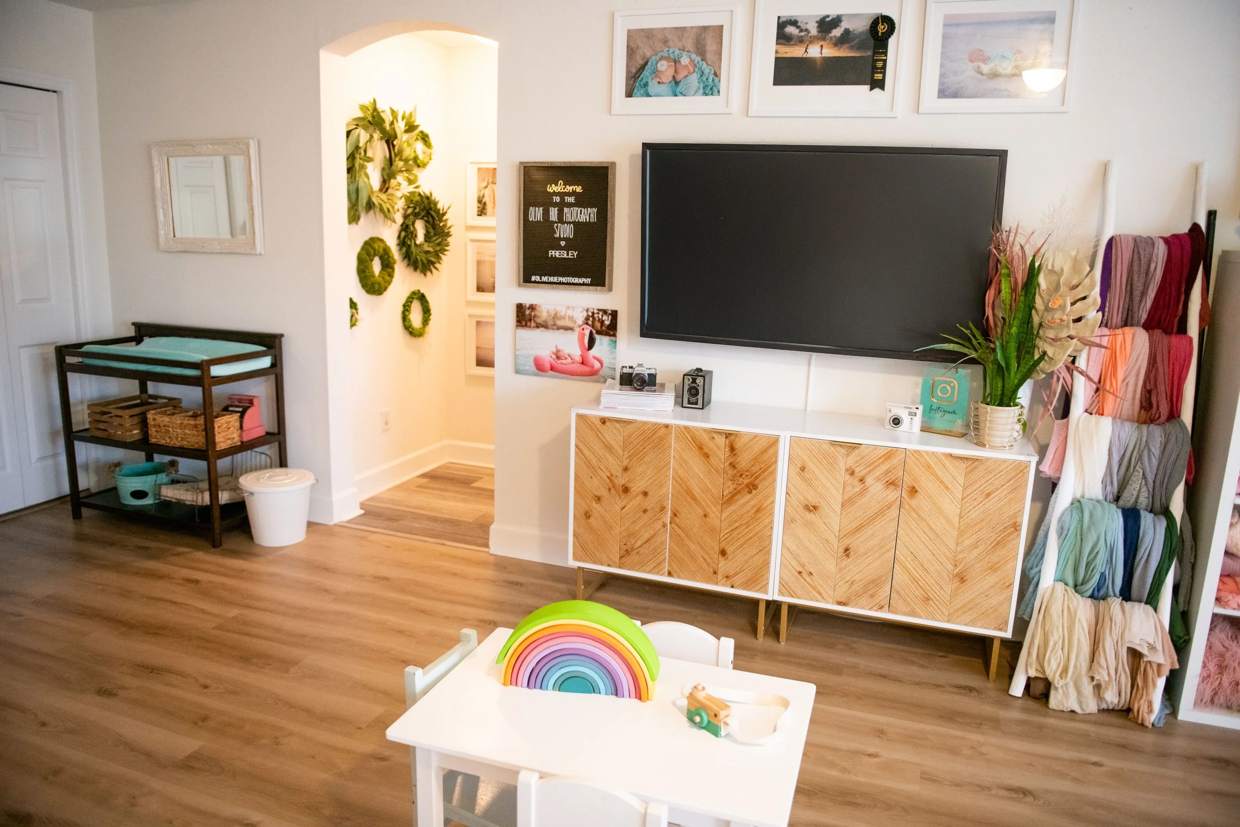 Living room corner with a white wall, mounted flat-screen TV, and a wooden sideboard. A small white table with a rainbow toy and a wooden toy train is in the foreground. A colorful towel rack with towels and scarves is on the right. A plant with pink
