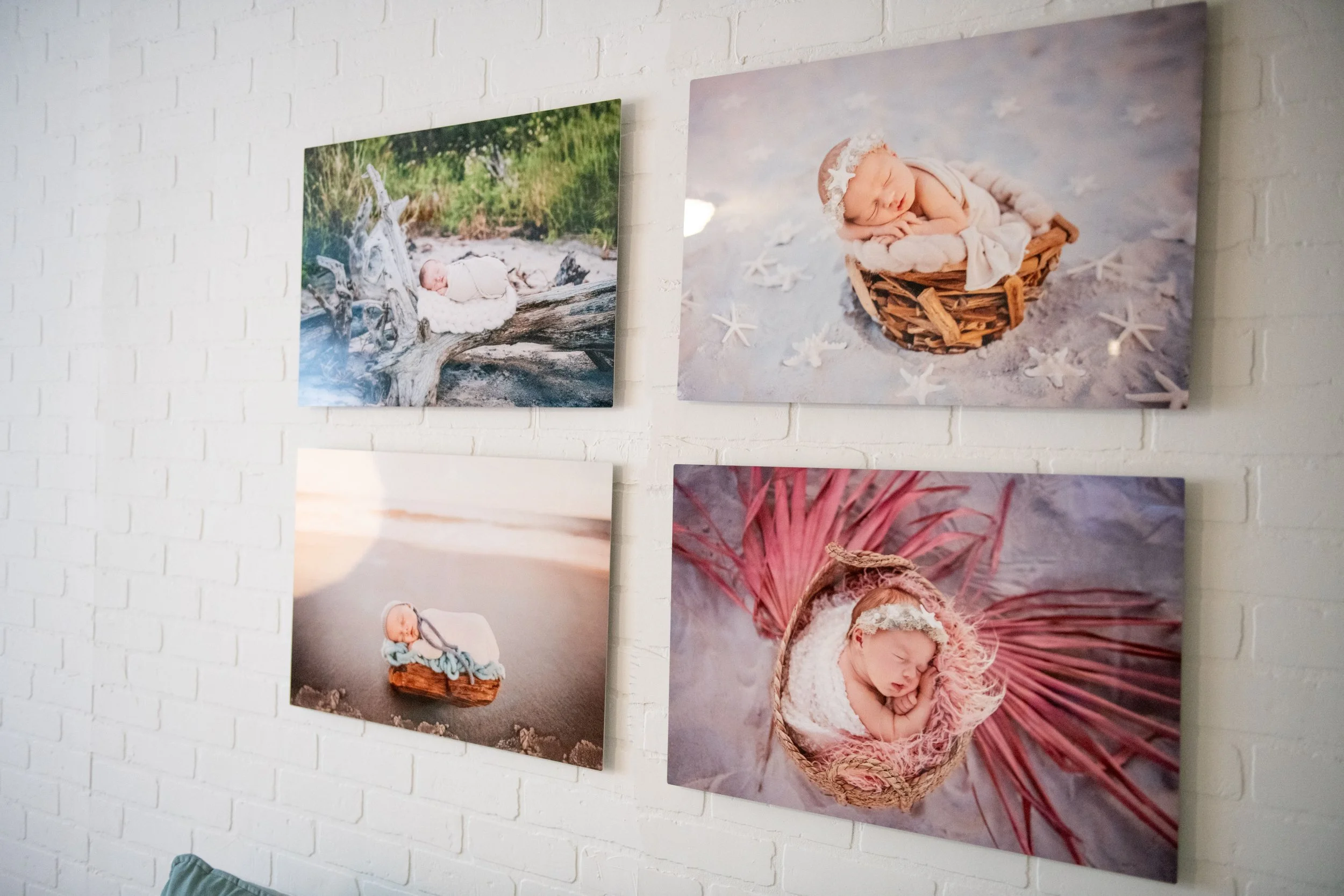 Four framed photographs of sleeping babies on a white brick wall, including a baby on a driftwood in a natural setting, a baby in a basket on a beach, a baby in a basket on a sandy surface with a sunset, and a baby in a basket on a pink leaf backgrou