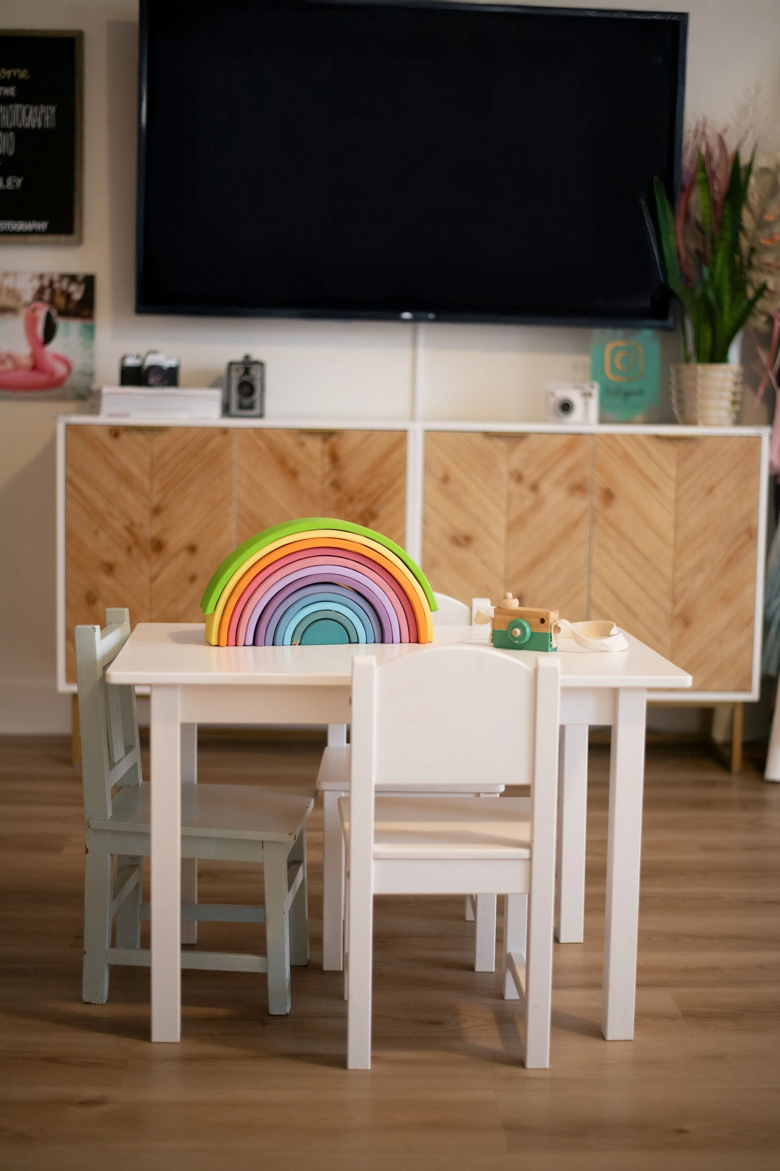 Small white children's table with three chairs, two are white and one is grey, with a colorful wooden rainbow toy on top. In the background, a wooden cabinet with a TV mounted above, a potted plant, and some decor including a camera and framed pictur