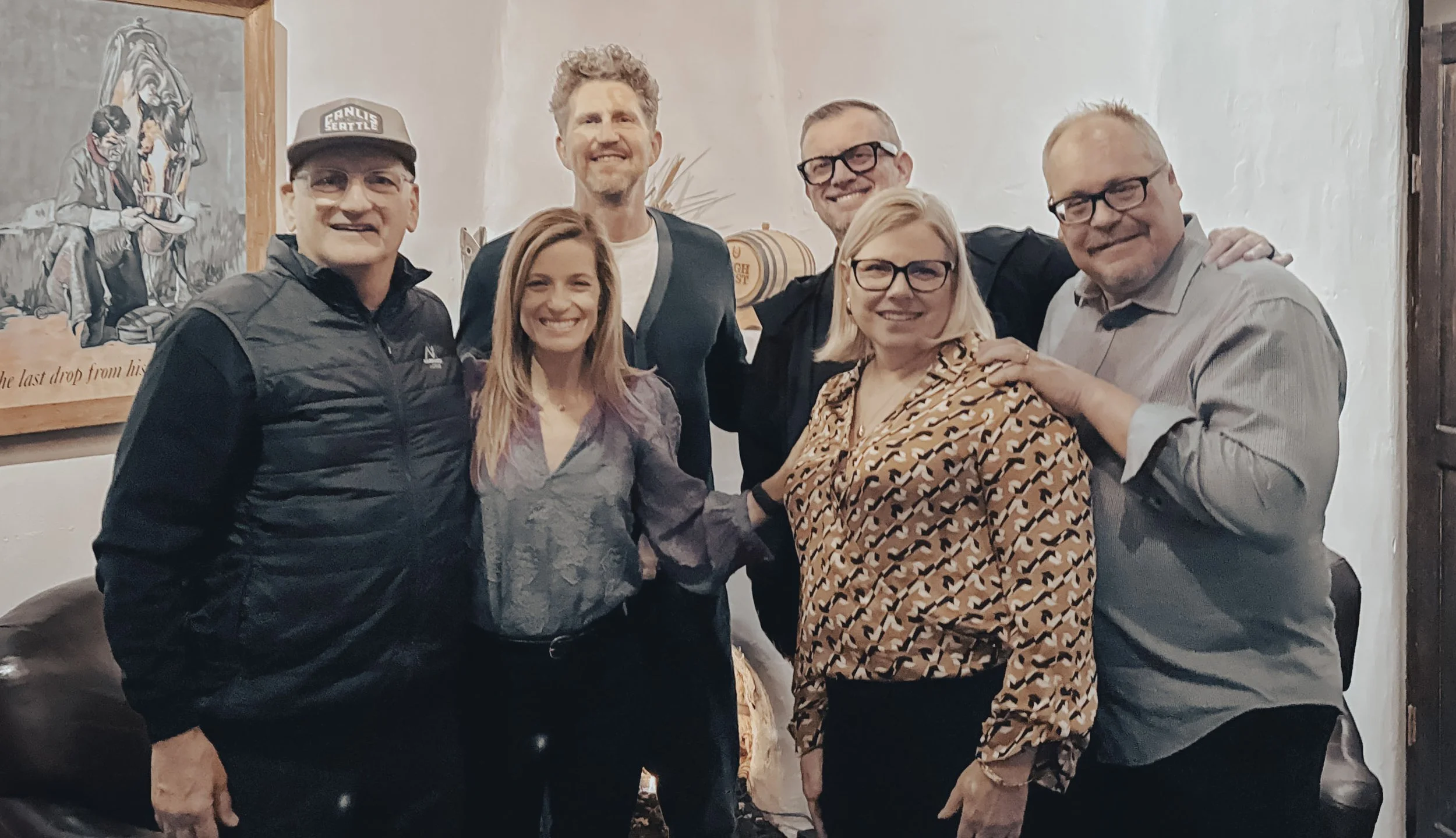 Group of six friends smiling and posing together indoors, with artwork and a vintage barrel in the background.
