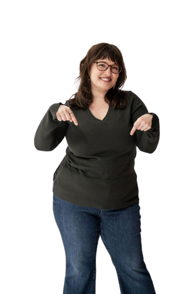 A woman with glasses and brown hair wearing a dark sweater and jeans, smiling and pointing down with both hands.