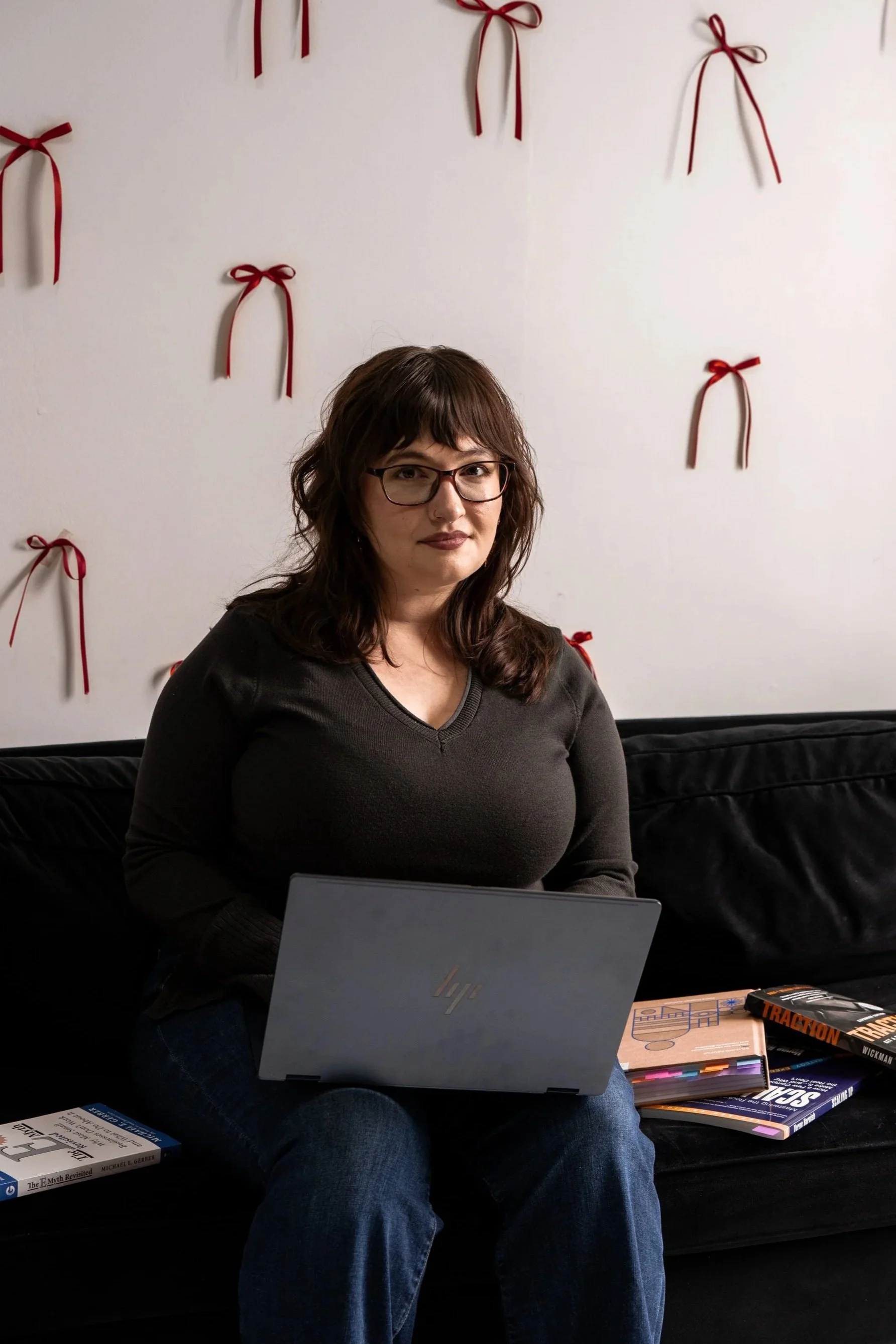 A woman with glasses sitting on a black couch with a laptop on her lap. Behind her is a wall decorated with red bows.