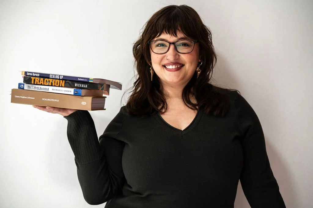 A woman with glasses and earrings smiling while holding a stack of books against a plain background.