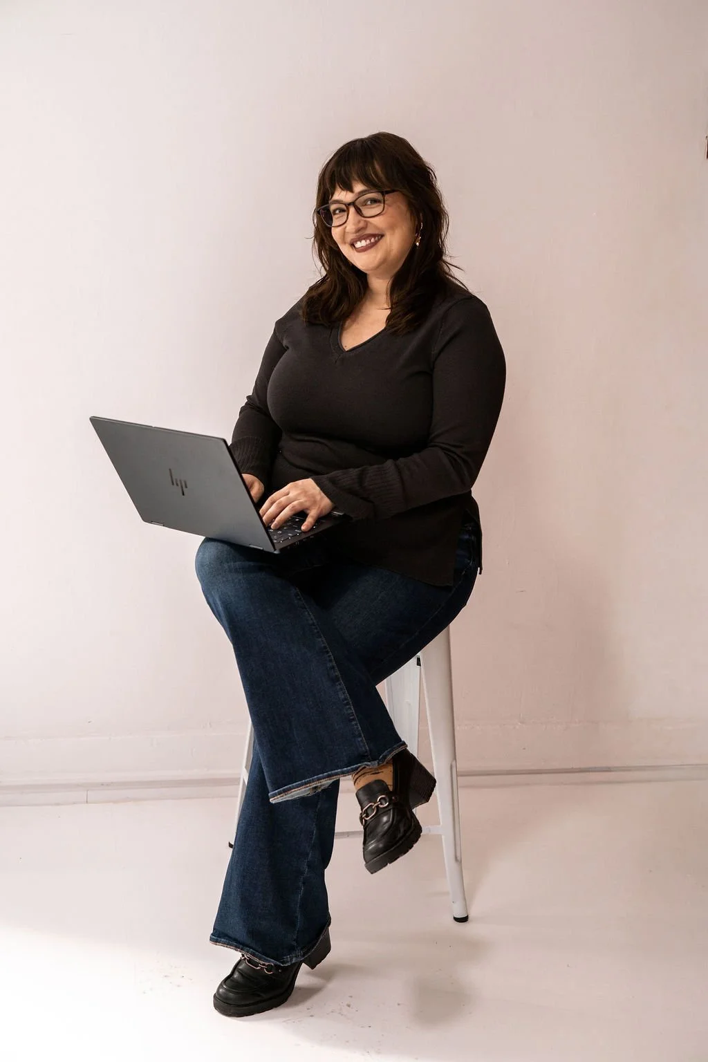 Woman sitting on a stool using a laptop in front of a plain wall.
