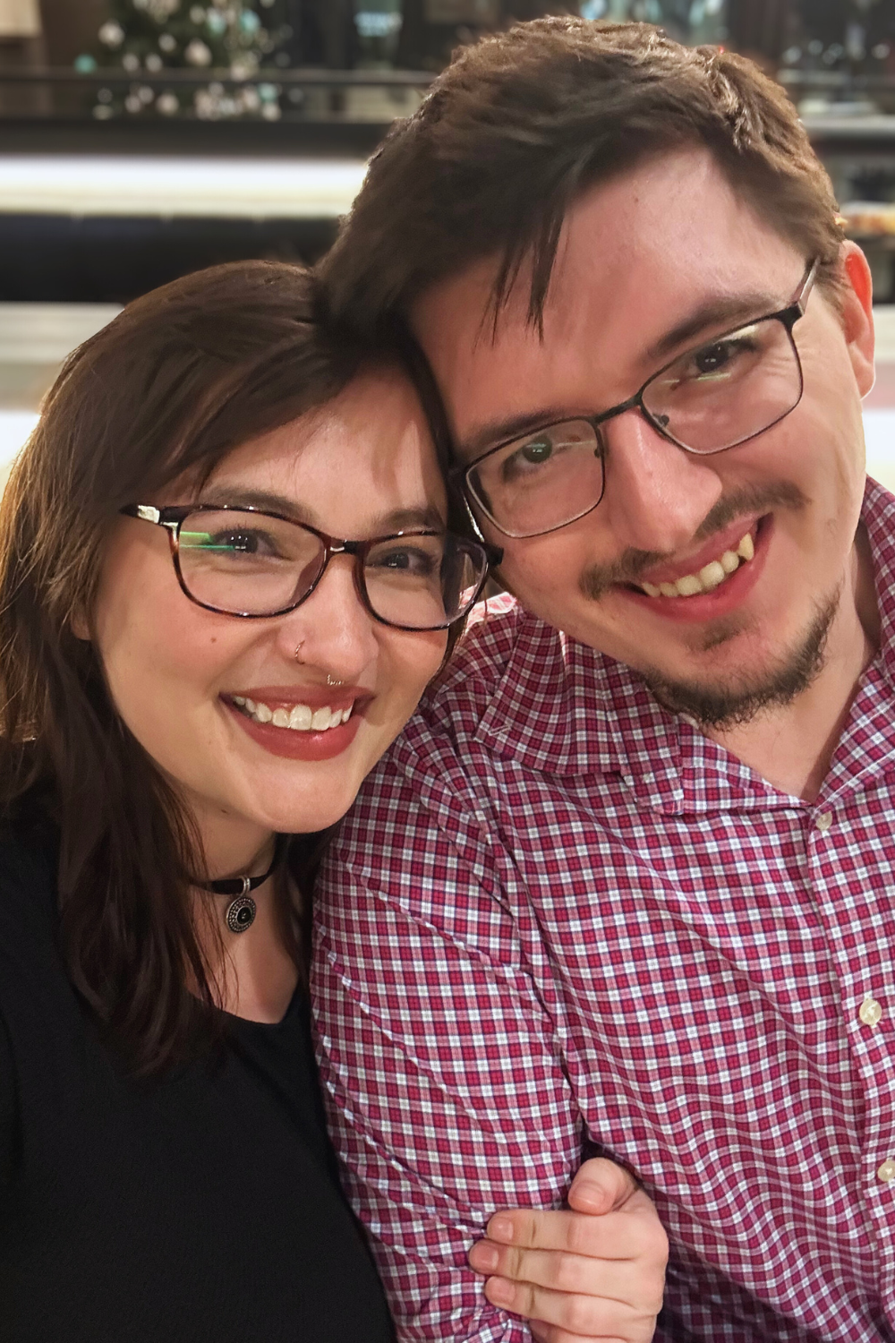 A smiling woman and man wearing glasses, close together, in a restaurant or cafe setting.