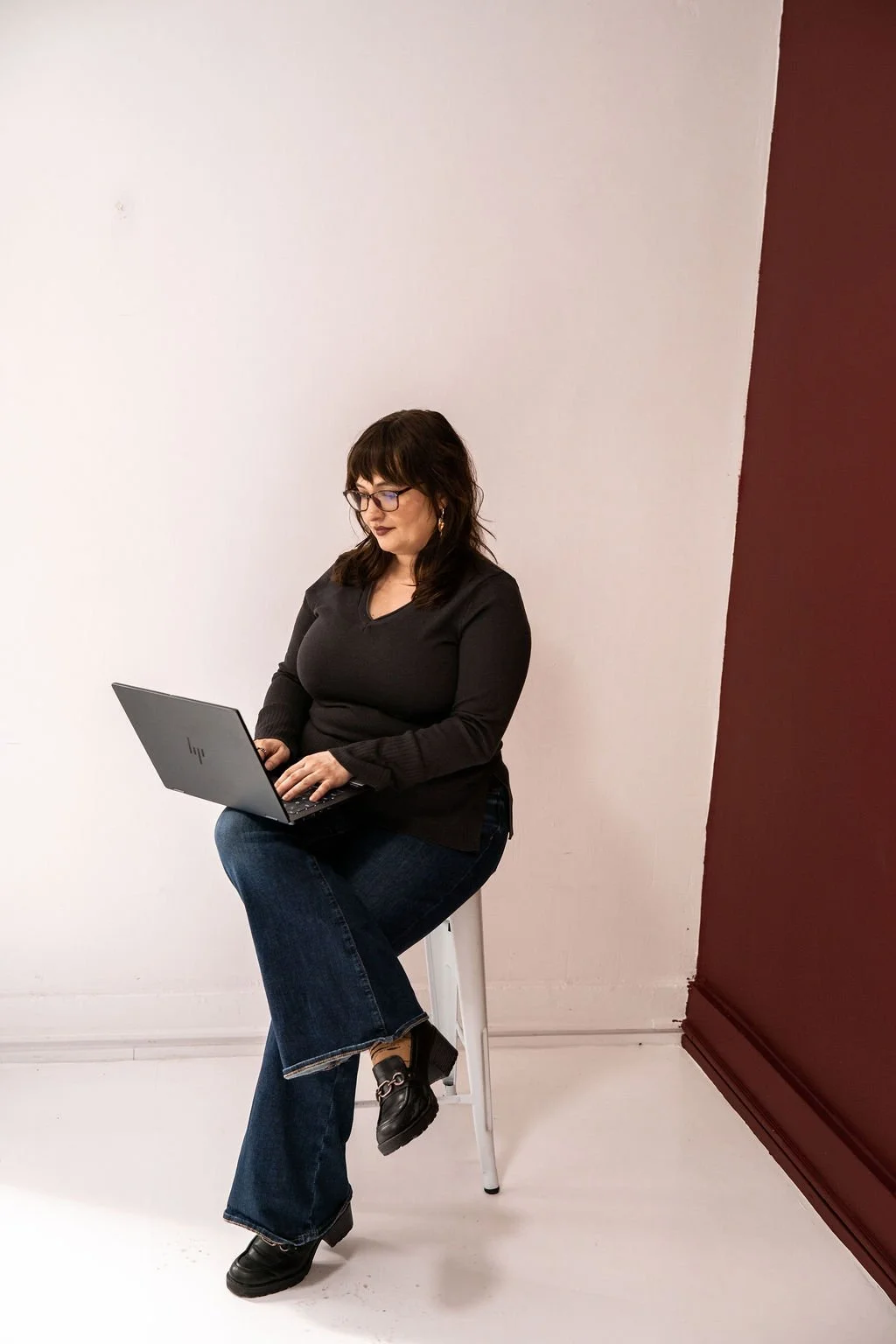 Woman with dark hair and glasses sitting on a stool, working on a laptop, against a plain white and maroon wall.