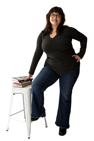 Woman with glasses and dark hair smiling, standing with one hand on her hip and the other resting on a small white stool with books, against a black background.