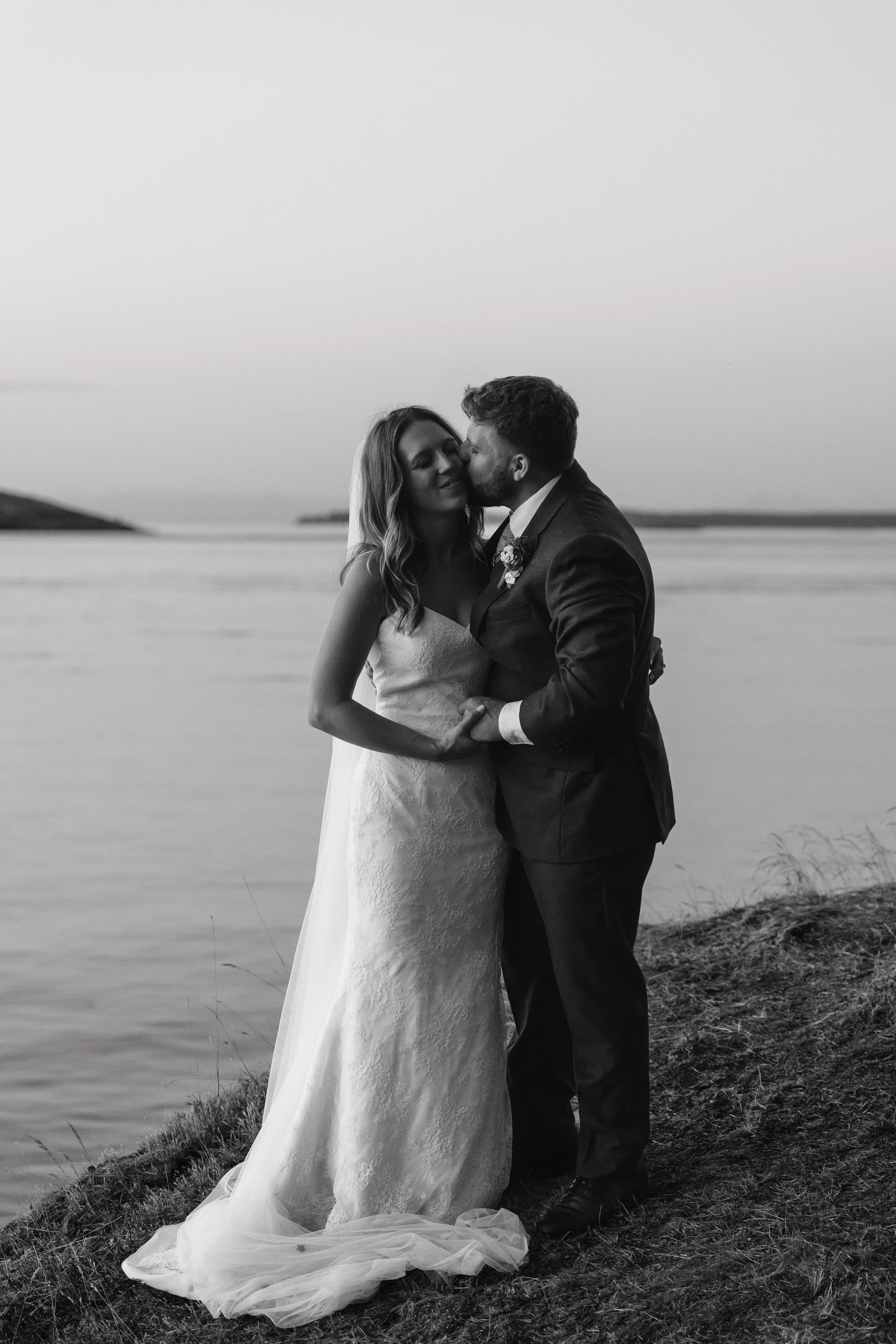 A black and white photo of a bride and groom embracing by a lake, kissing, with a natural landscape in the background.