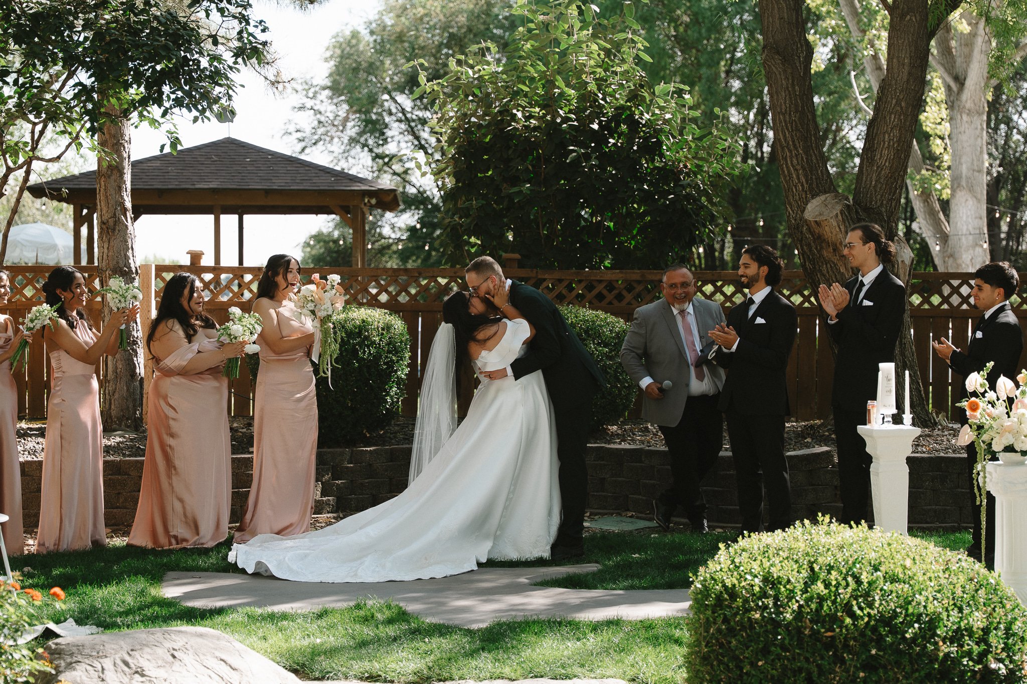 A couple in wedding attire sharing a kiss during an outdoor wedding ceremony, surrounded by bridesmaids holding bouquets, groomsmen, and a cheerful officiant, under trees and a wooden gazebo.