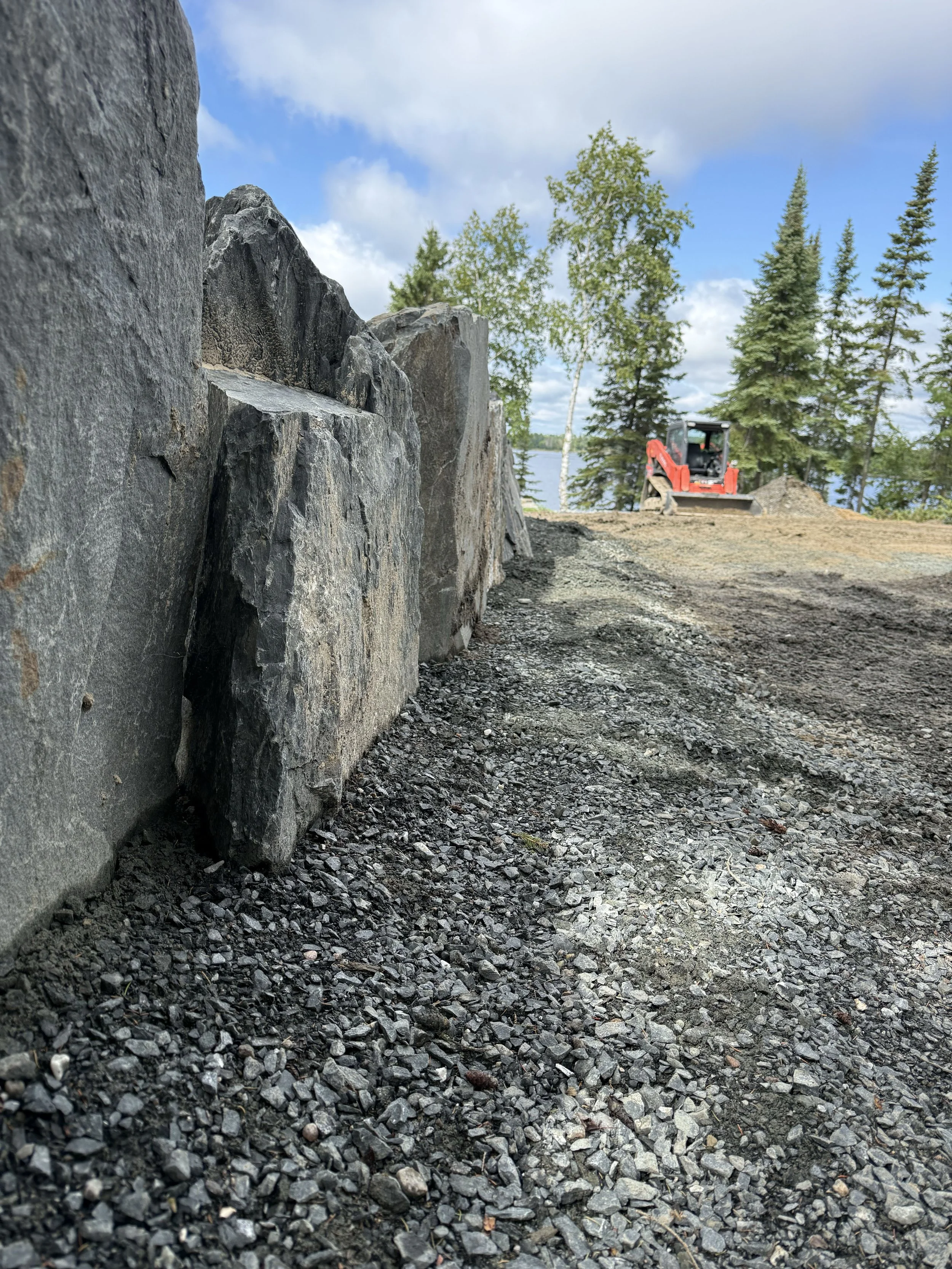 Construction site where large rocks are being placed along the edge of a gravel path near trees and a body of water. A red compact excavator is visible in the background.
