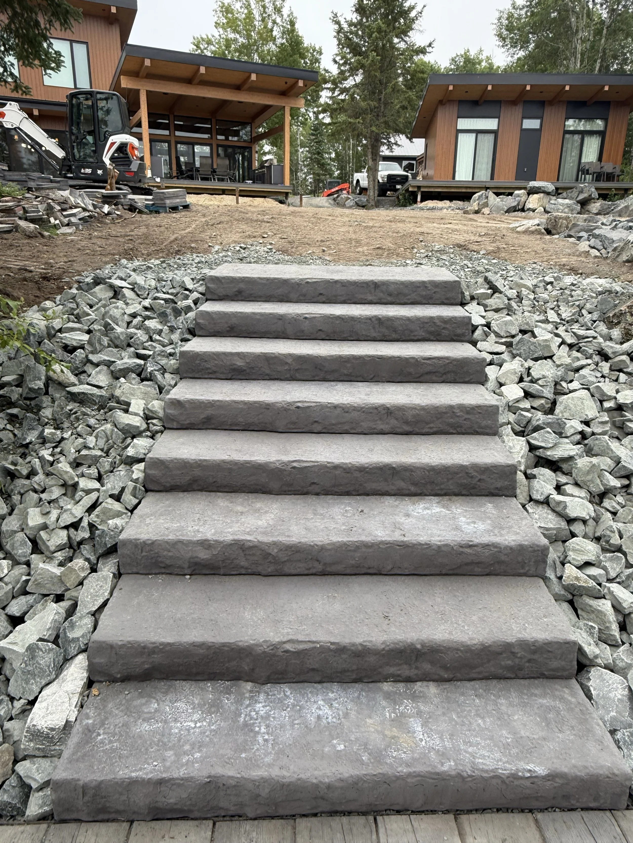 Concrete stairs leading up to a dirt pathway in front of modern houses with wooden siding, surrounded by trees and construction equipment.