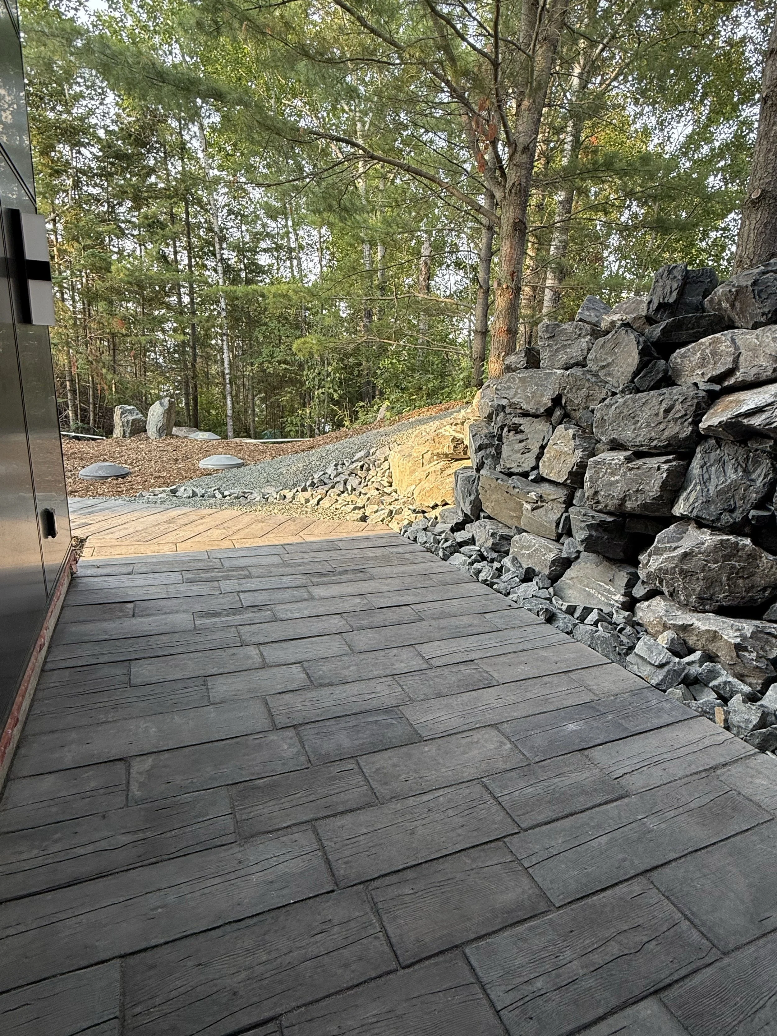 Outdoor patio with gray stone-like tiles, bordered by a stacked dark gray stone wall, with a wooded area and sunlight filtering through trees in the background.
