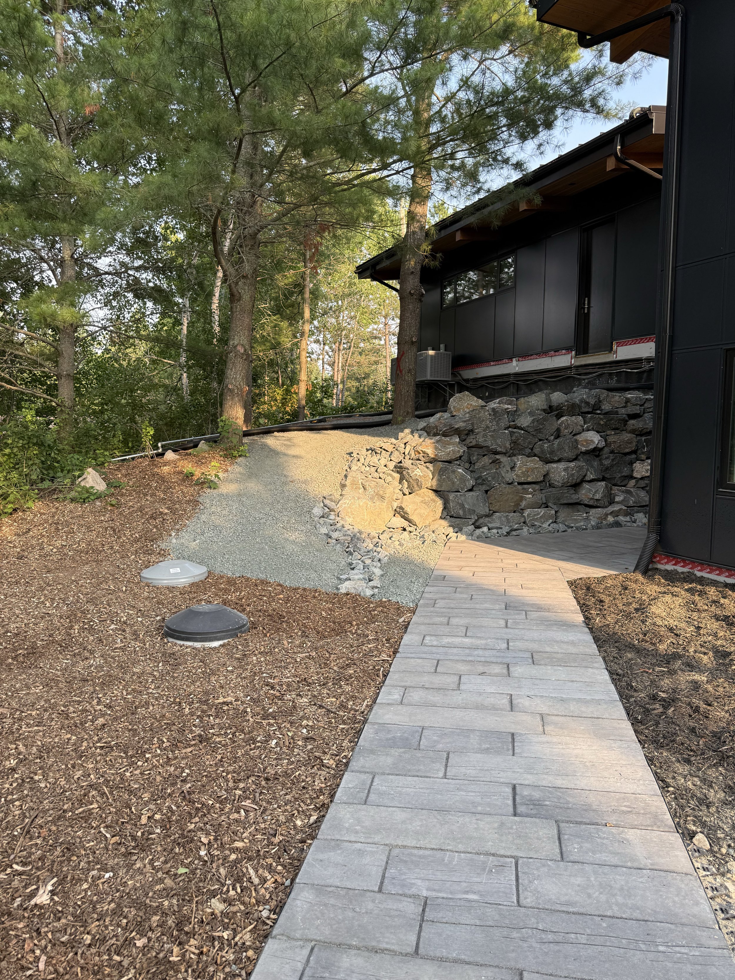 Part of a newly paved sidewalk leading to a house with black siding, surrounded by trees and rocks, with construction materials present.