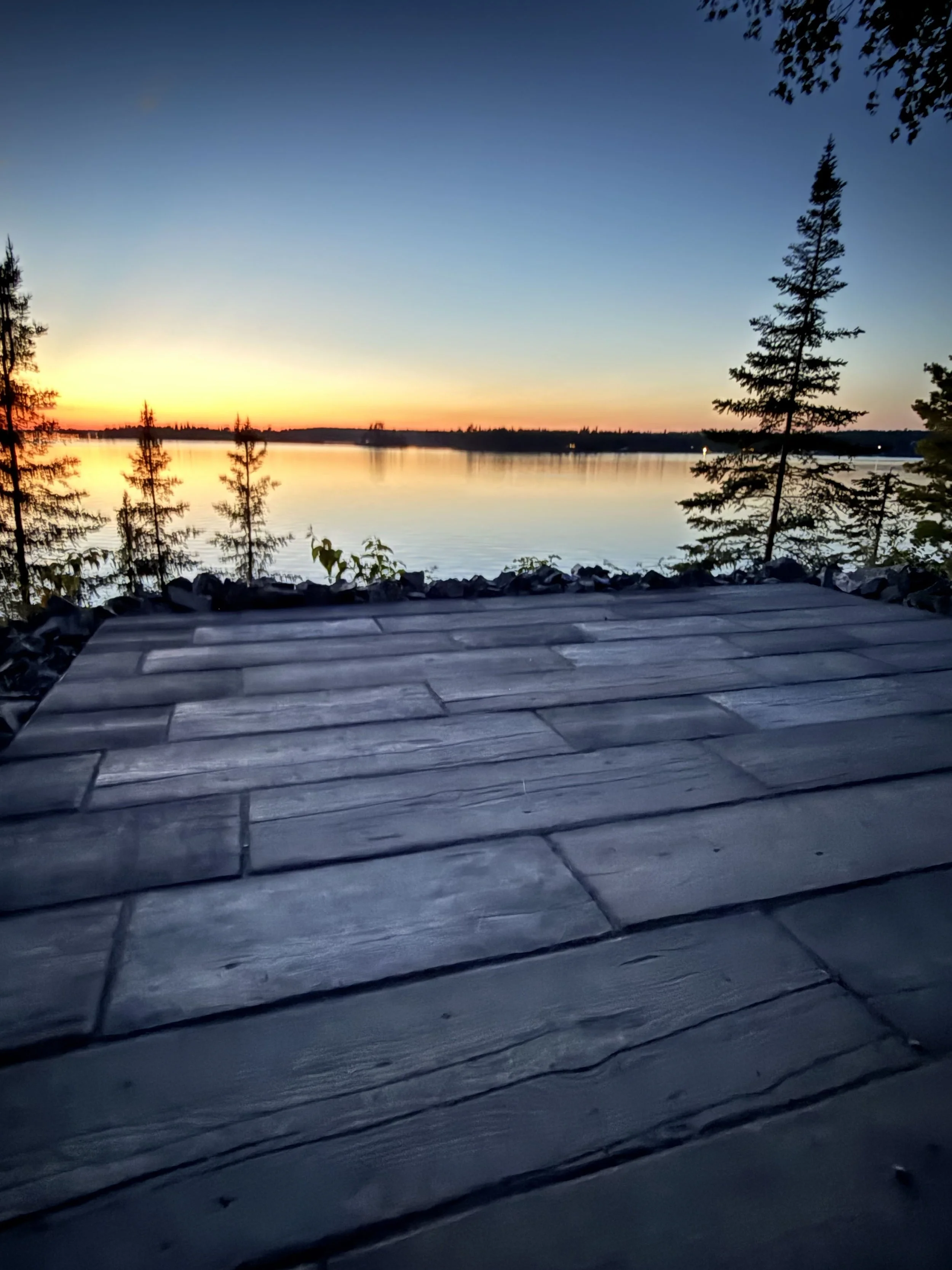 View of a calm lake at sunset, with trees on the shore and a tiled surface in the foreground.