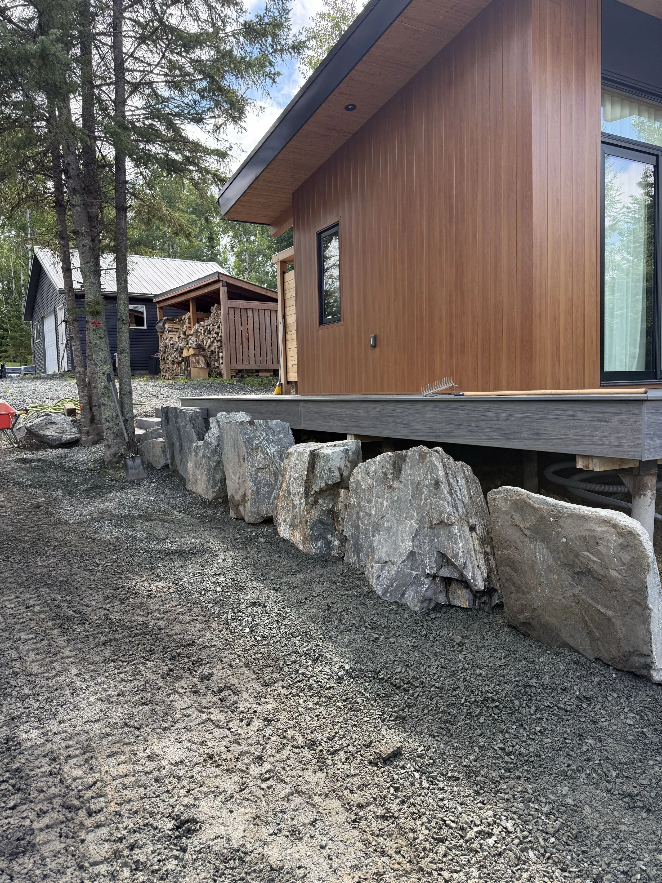 Modern house with a wooden exterior, set on a foundation with large rocks along the edge, surrounded by trees.