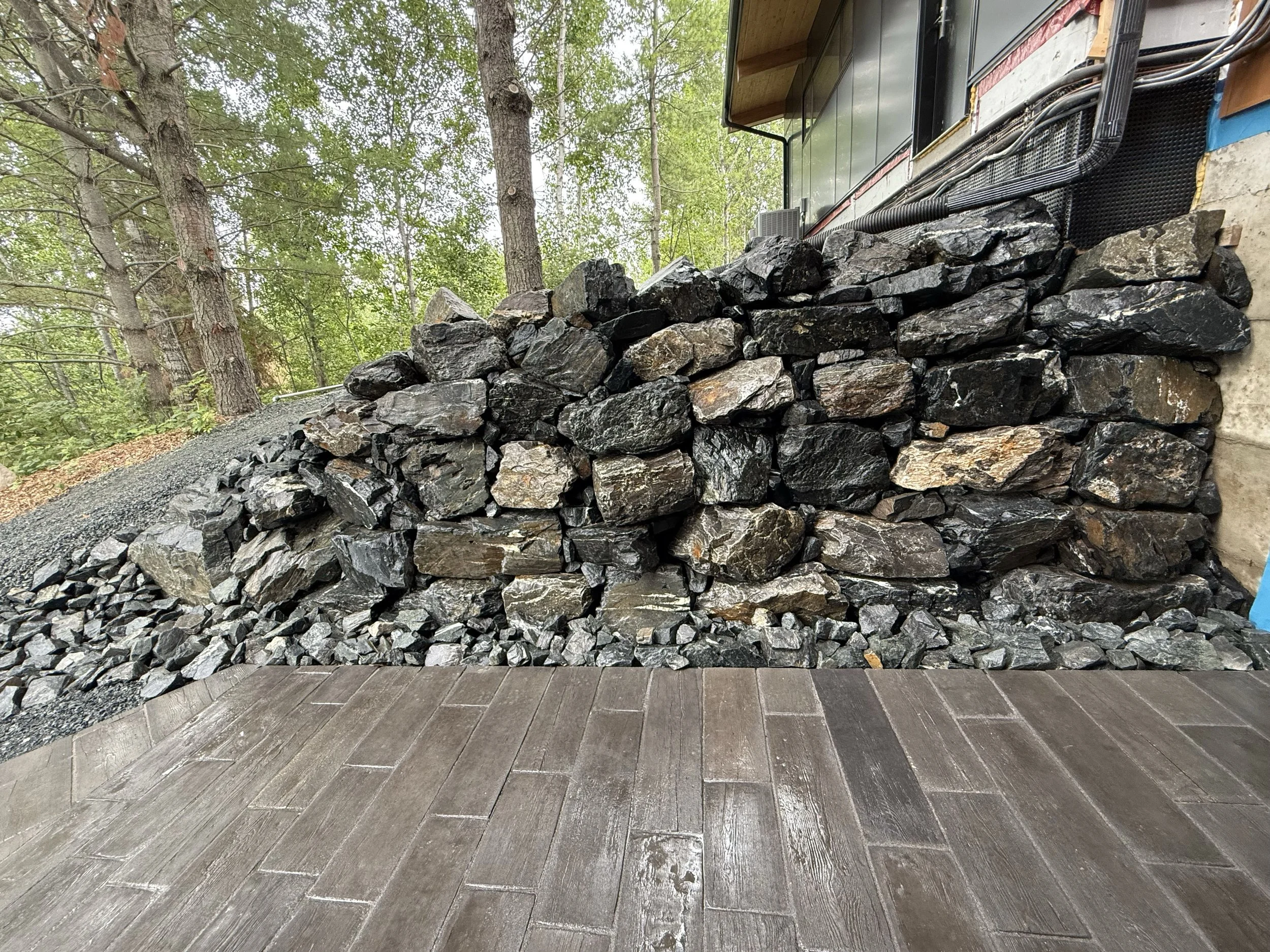 A large pile of darkly-colored natural rocks stacked against the side of a house, with trees and greenery in the background and a wooden deck in the foreground.