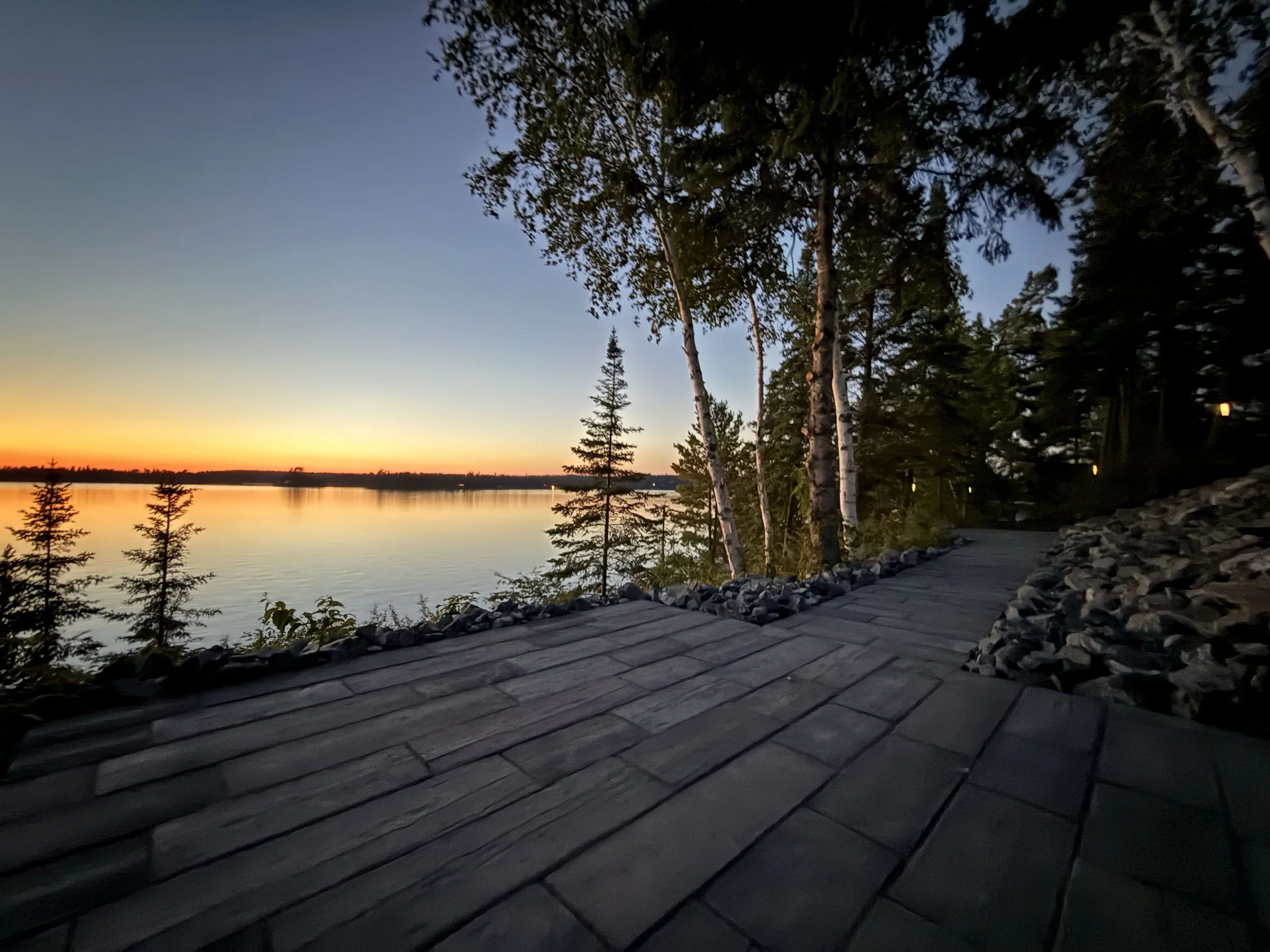 Sunset over a calm lake viewed from a wooden pathway through a forest with tall trees.