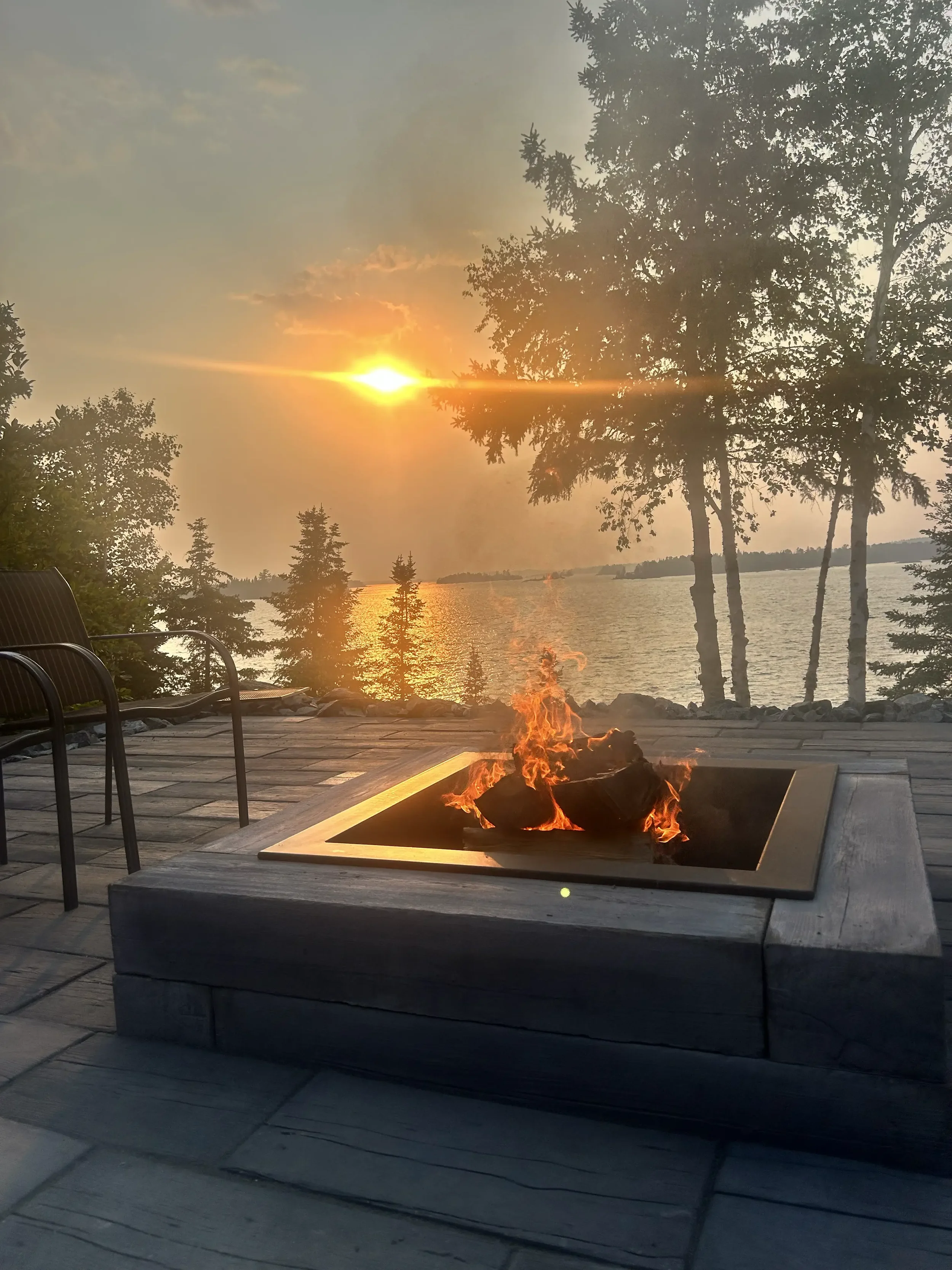 Sunset over a body of water with trees in the foreground, and a small fire pit on a patio in the foreground.