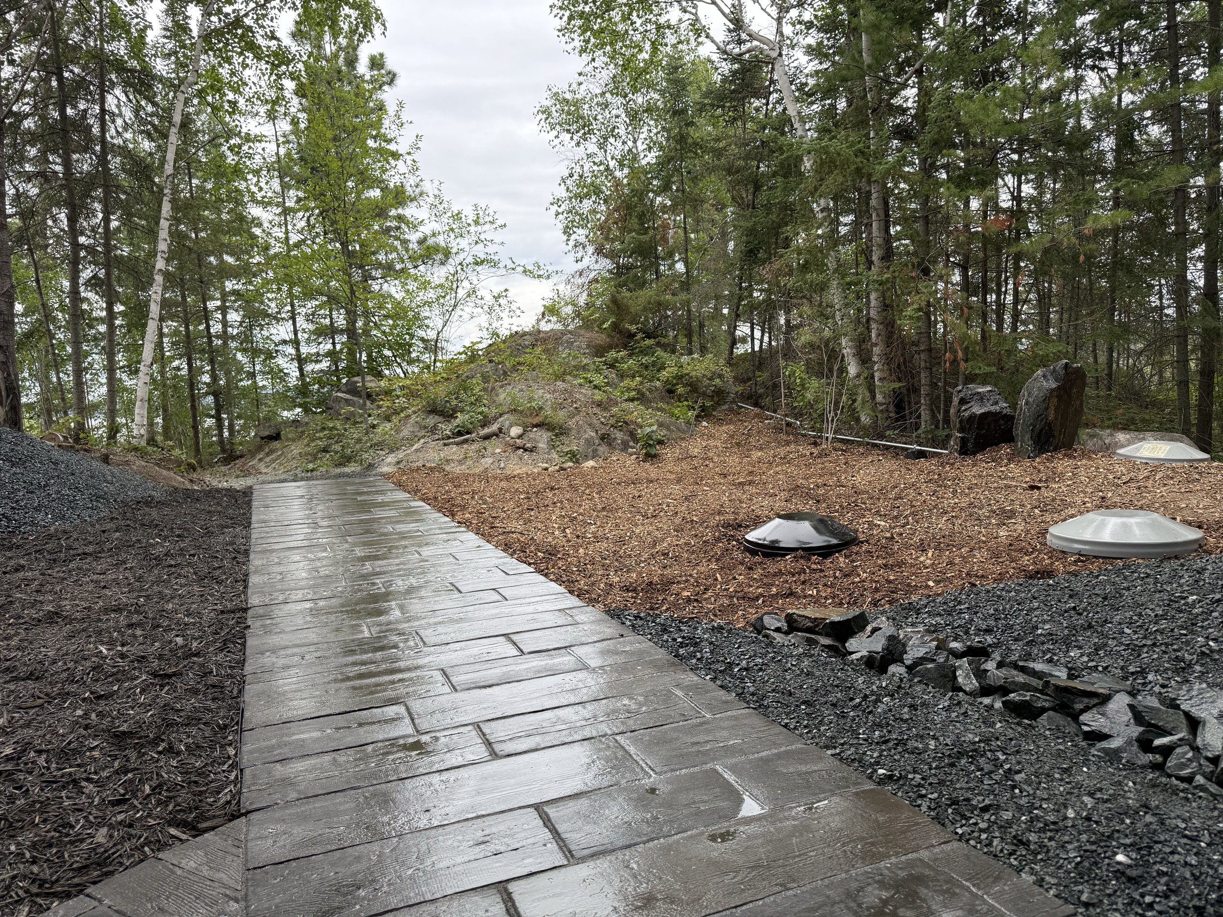 Wet paved walkway goes through a wooded area with trees and rocks, with underground utility covers visible on the right side.
