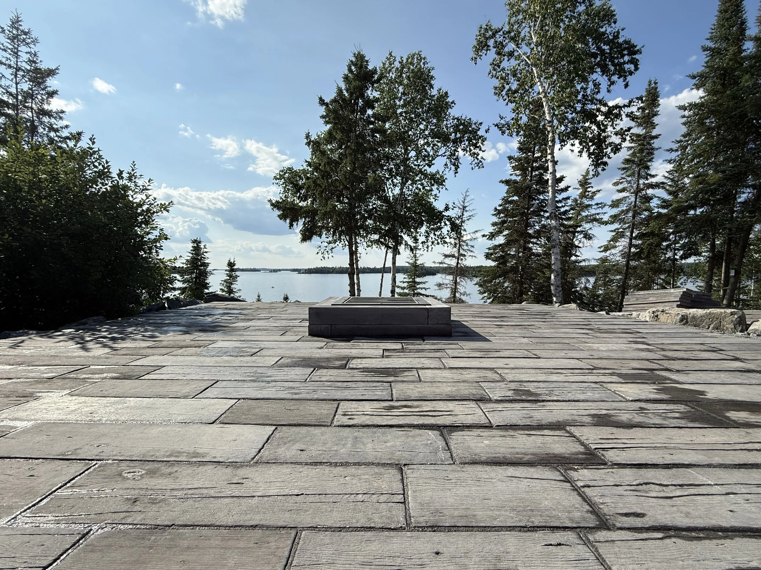 View from a stone patio overlooking a lake, surrounded by trees and with a partly cloudy sky.