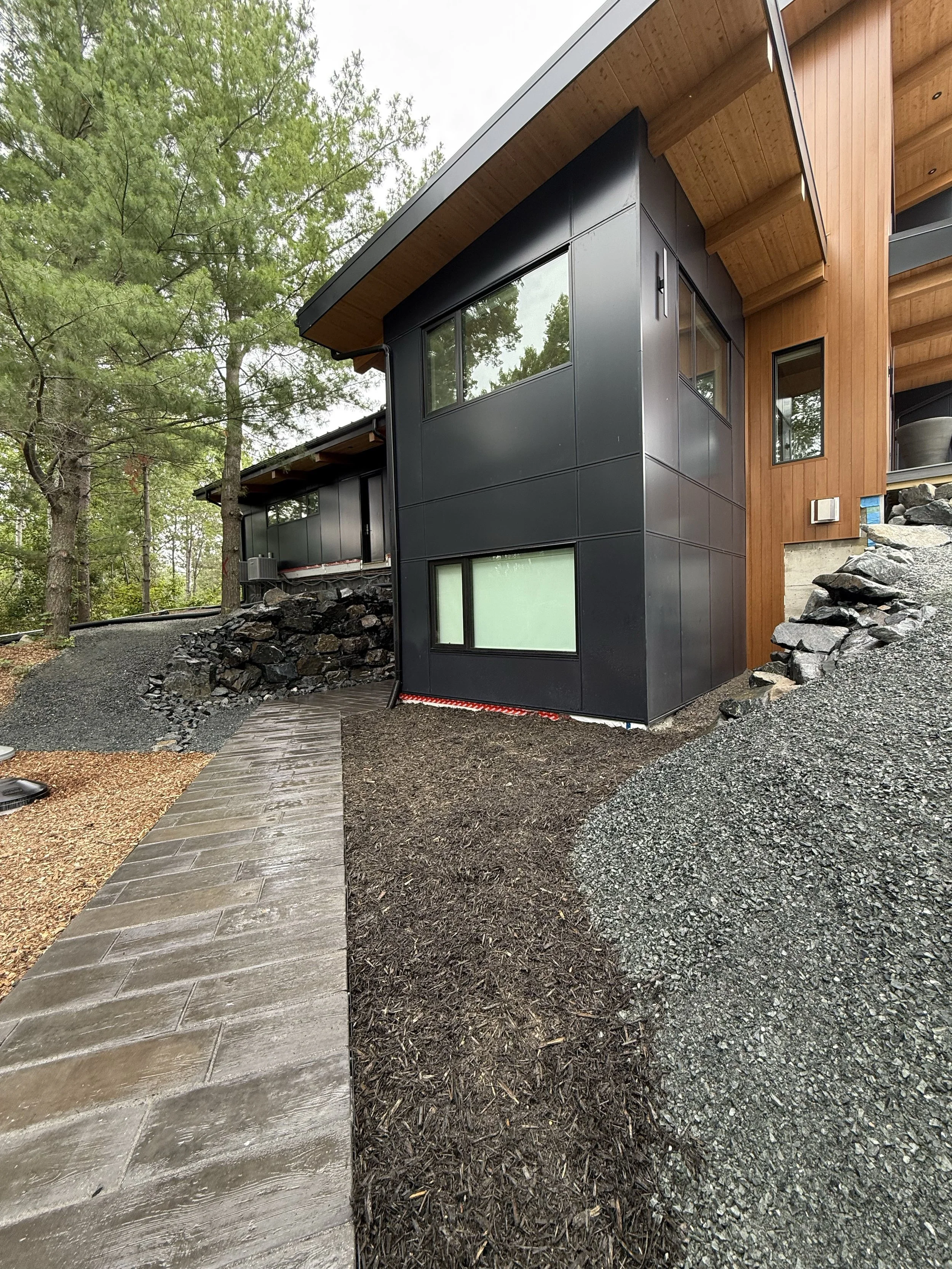 Modern house exterior with black and wooden siding, large windows, surrounded by trees and landscaping.