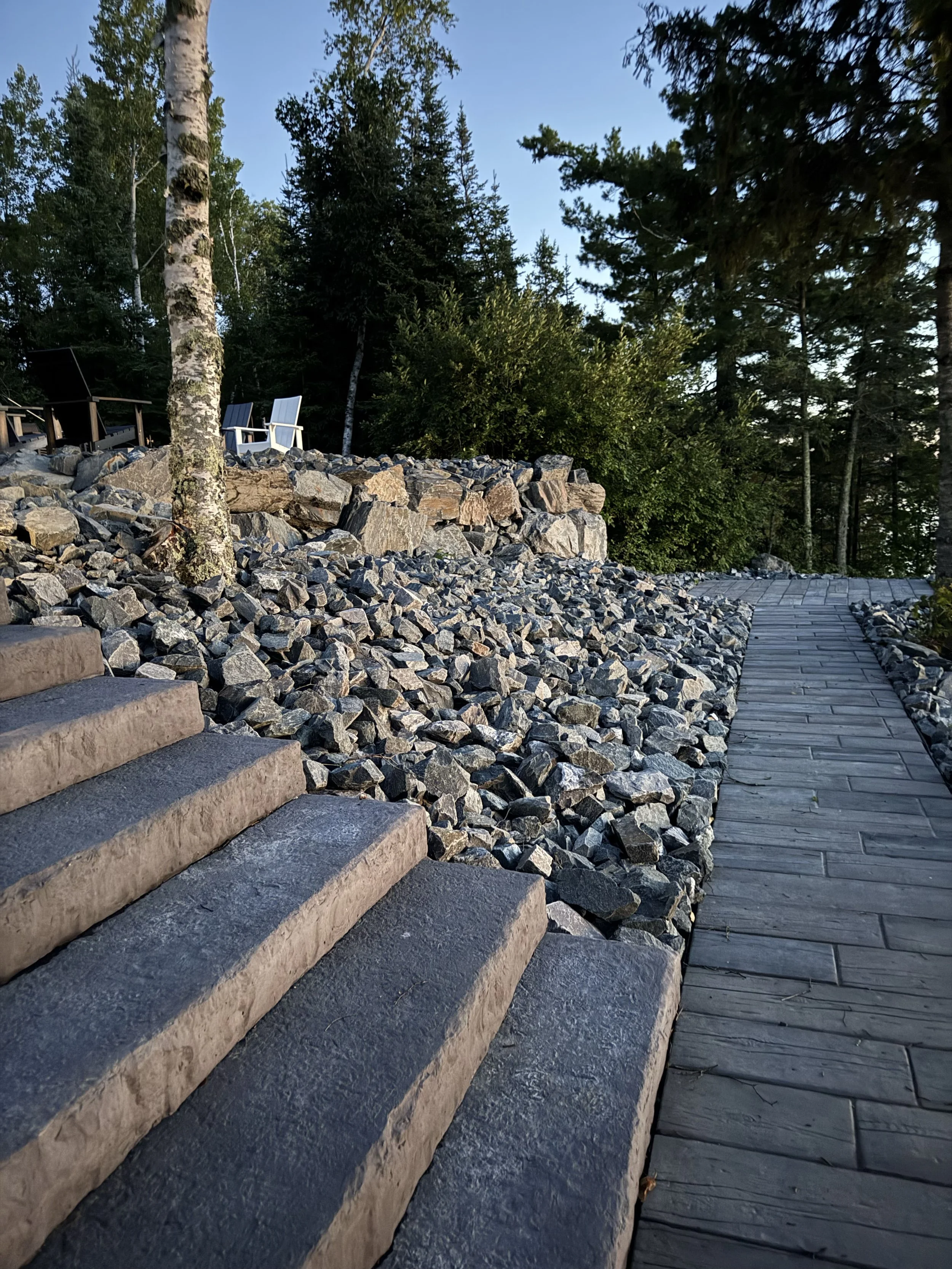 A landscaped outdoor area with stone steps on the left and a wooden pathway on the right, surrounded by rocks and trees.