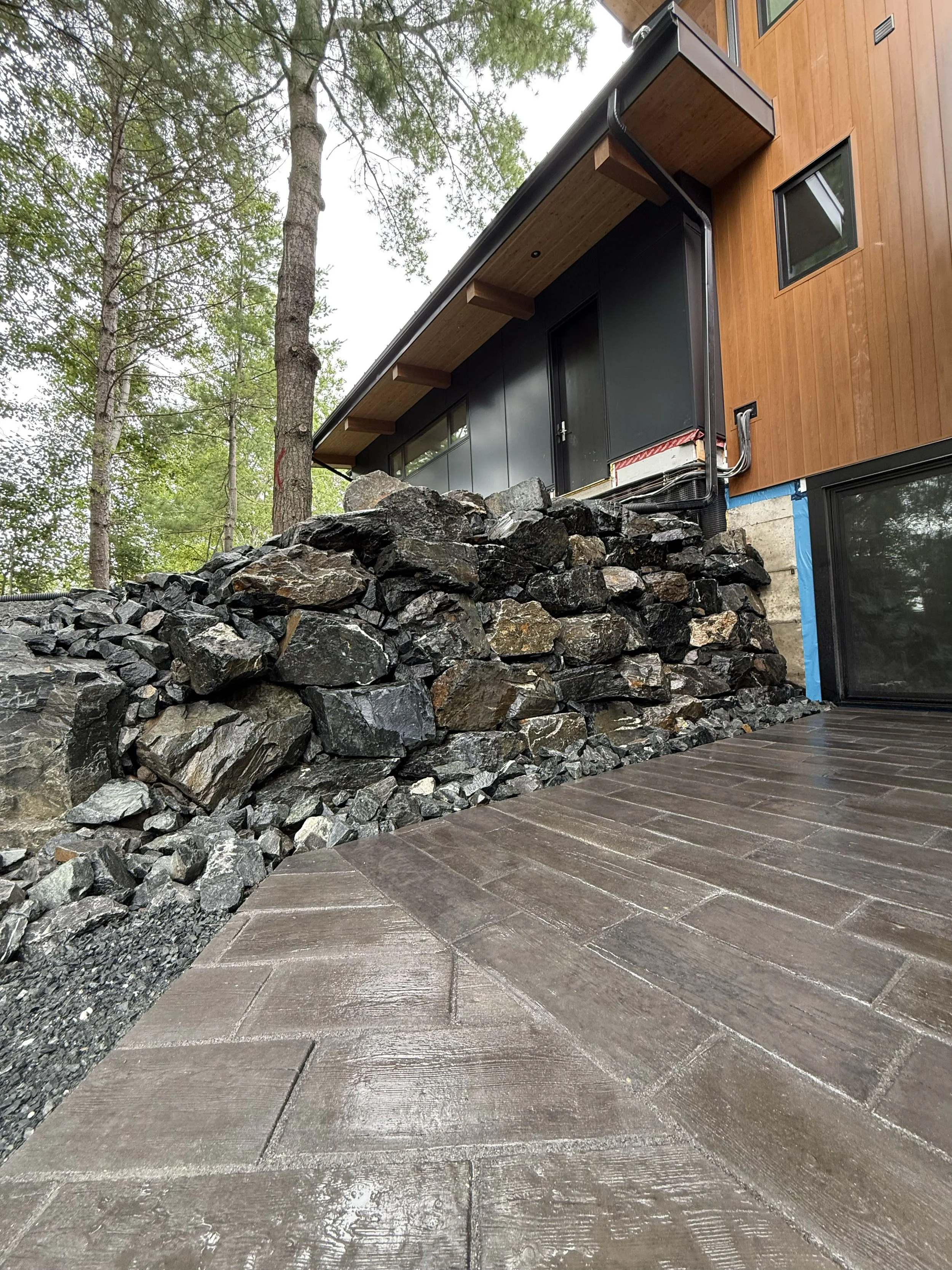 Close-up view of a modern house with a black exterior, wooden accents, and a stone retaining wall, situated in a wooded area with tall trees.
